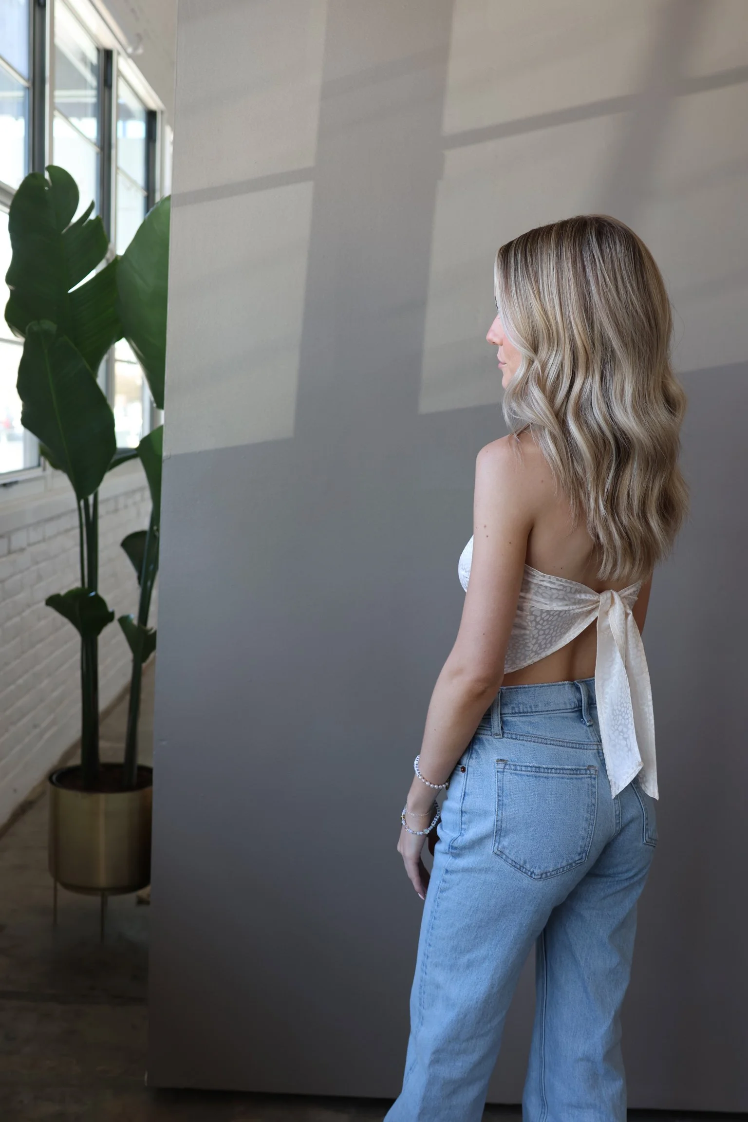 Side view of a woman with wavy blonde hair wearing a white, backless top and high-waisted blue jeans, standing indoors near a large window and a gray wall.