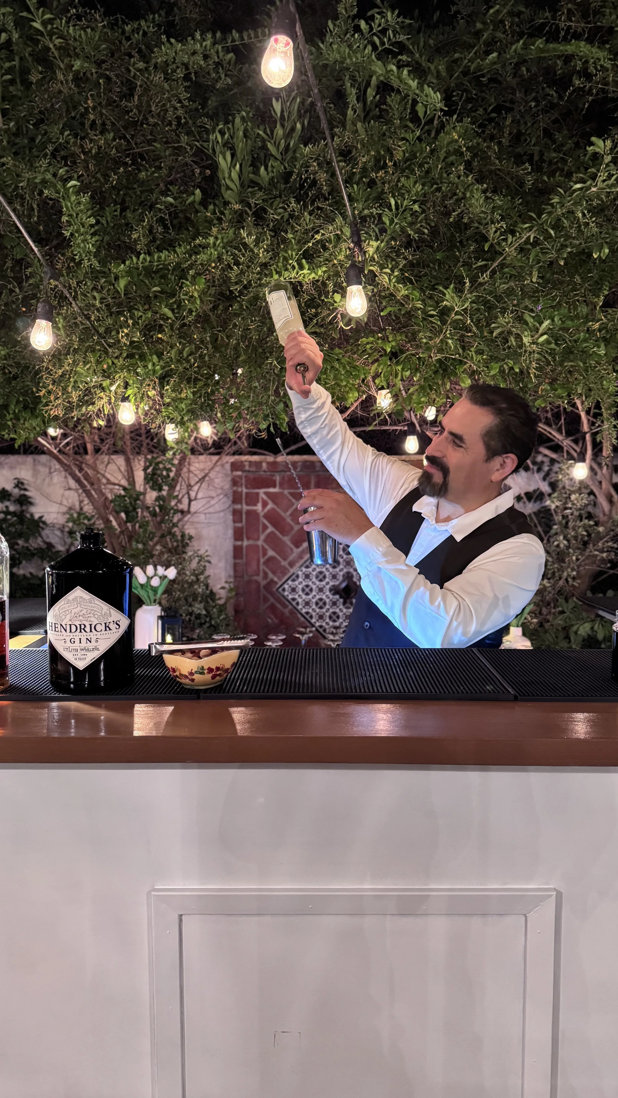 A bartender in a white shirt and black vest pouring a drink from a shaker behind an outdoor bar with string lights overhead, greenery in the background, and a bottle of Hendrick's gin on the bar.