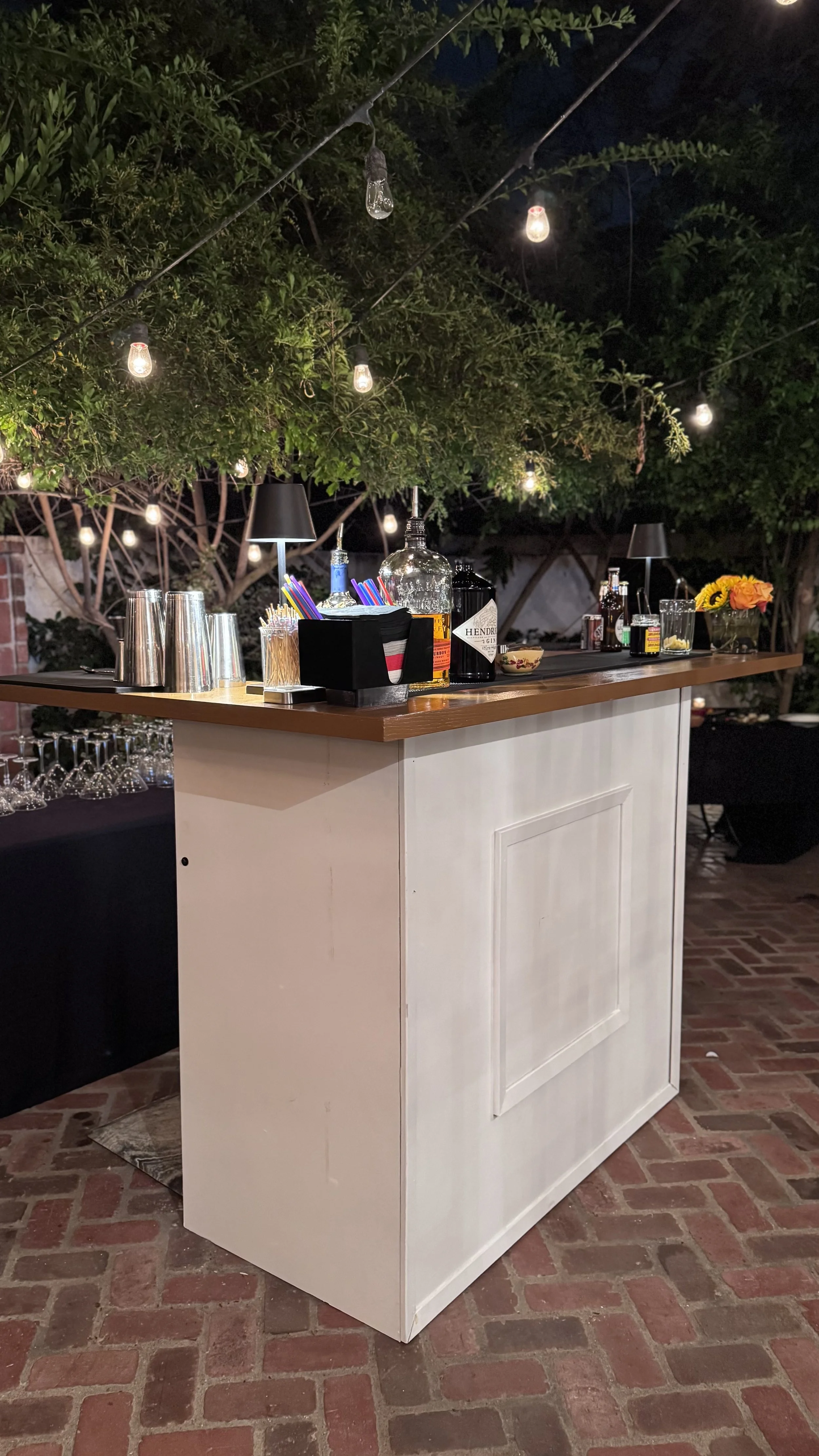 Outdoor bar setup with liquor bottles, cocktail glasses, and bar tools on a wooden counter. String lights hang above, and trees or bushes are in the background.