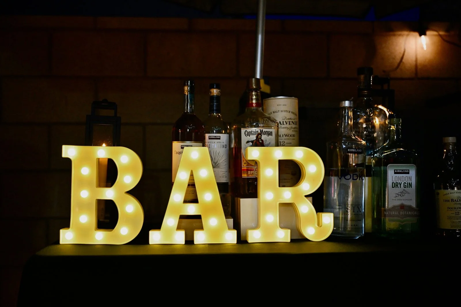 Illuminated sign spelling 'BAR' with letter bulbs, placed in front of bottles of alcohol and mixers on a bar counter, in a dimly lit setting.
