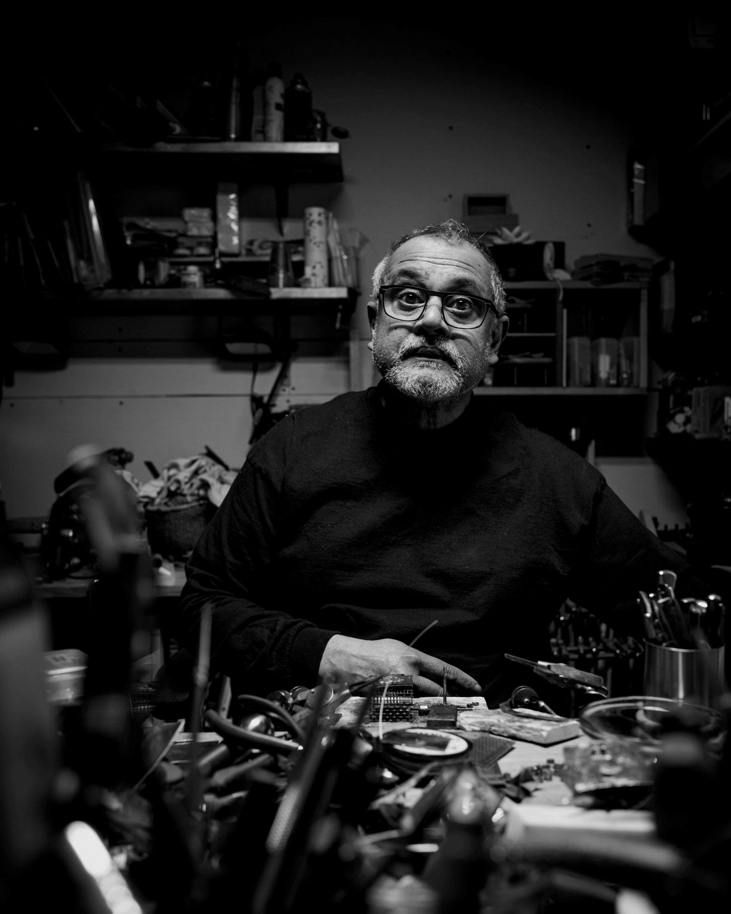 A black and white photo of a man with glasses, a beard, and gray hair, sitting at a cluttered workbench in a workshop, surrounded by tools and supplies, staring at the camera.