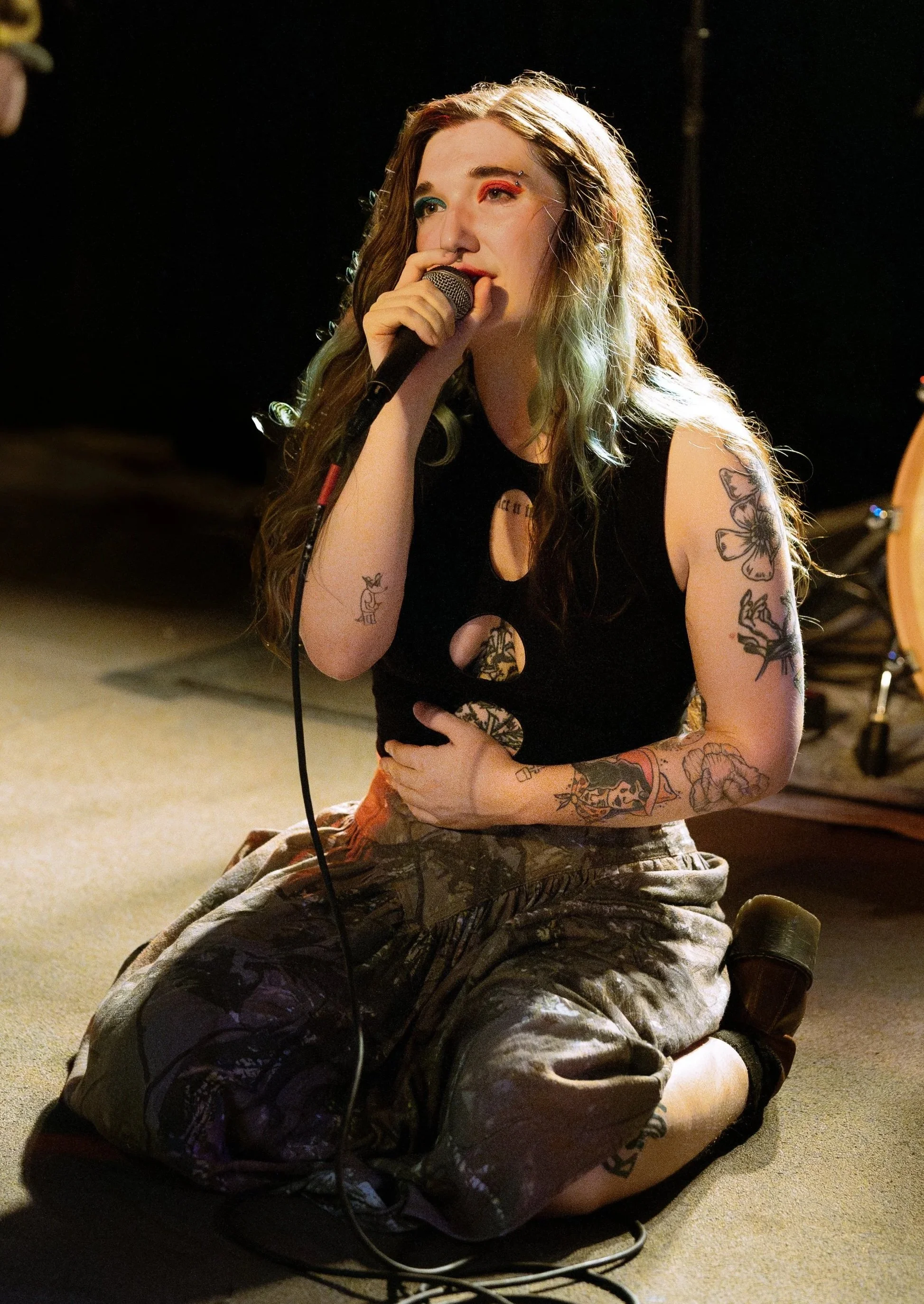 A woman with wavy hair and tattoos, sitting on her knees on the floor, singing into a microphone in a dimly lit music studio or stage with drums in the background.