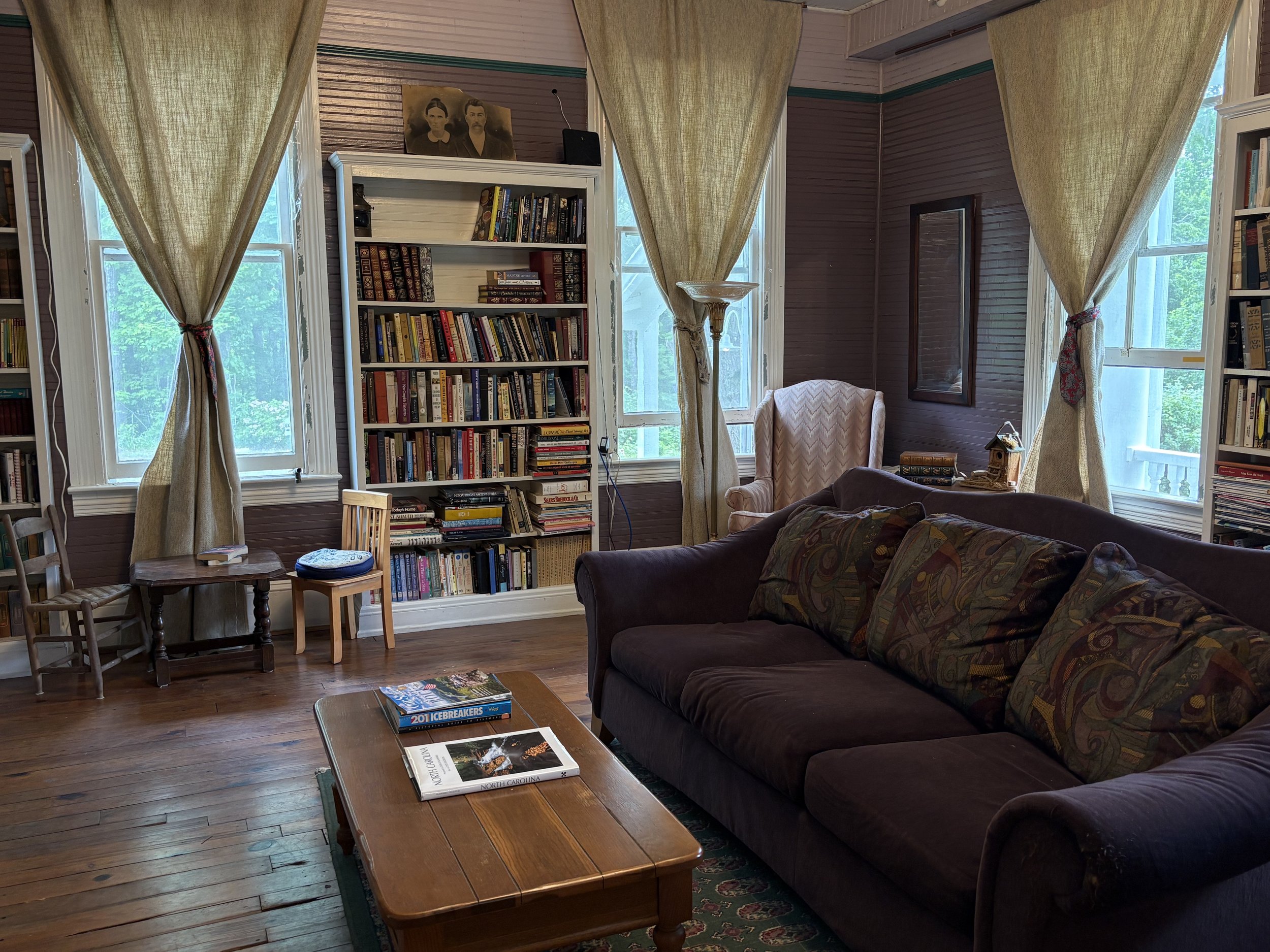 A young boy sitting in a pink upholstered armchair reading a book in a cozy living room with wooden floors, a patterned area rug, a tall bookshelf filled with books, a window, and a floor lamp.
