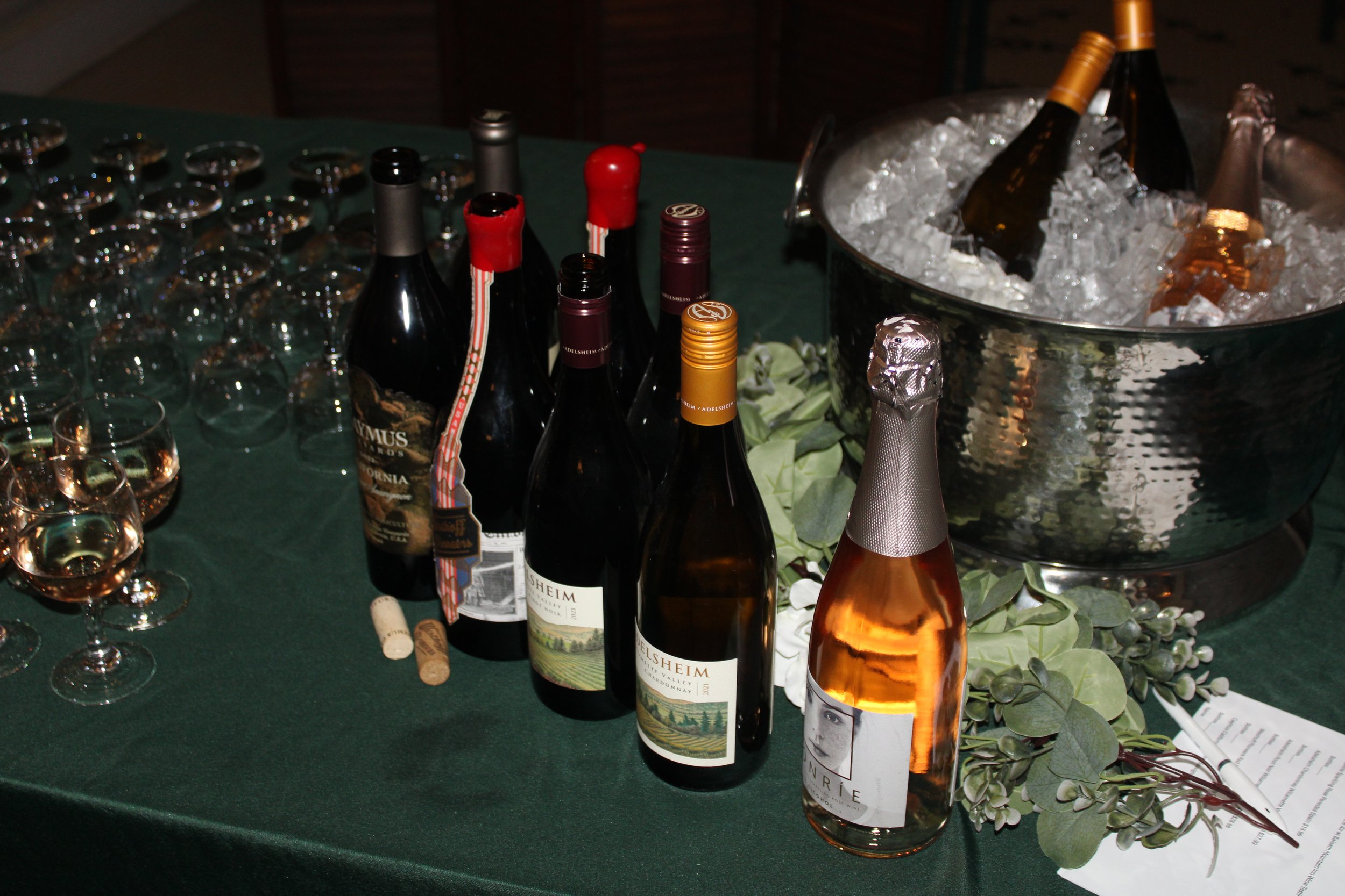 A small gathering of people at a wine-tasting event inside a well-lit room with large windows. There is a table with empty wine glasses and bottles. A woman is serving wine, and several guests are chatting and tasting. A beverage refrigerator is in the corner.