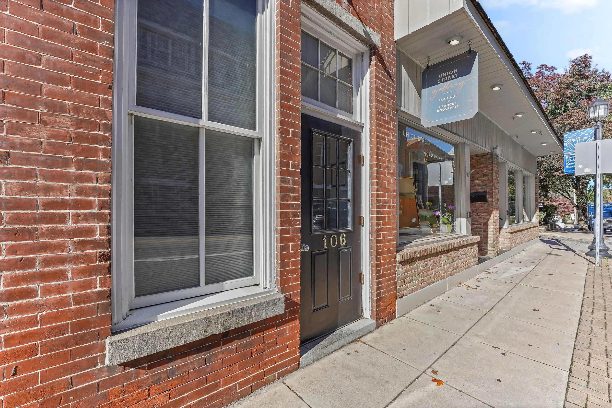 Exterior view of a mixed-use brick building in Kennett Square of Chester County PA. The sidewalk is on the right with a blue street sign and trees in the background.