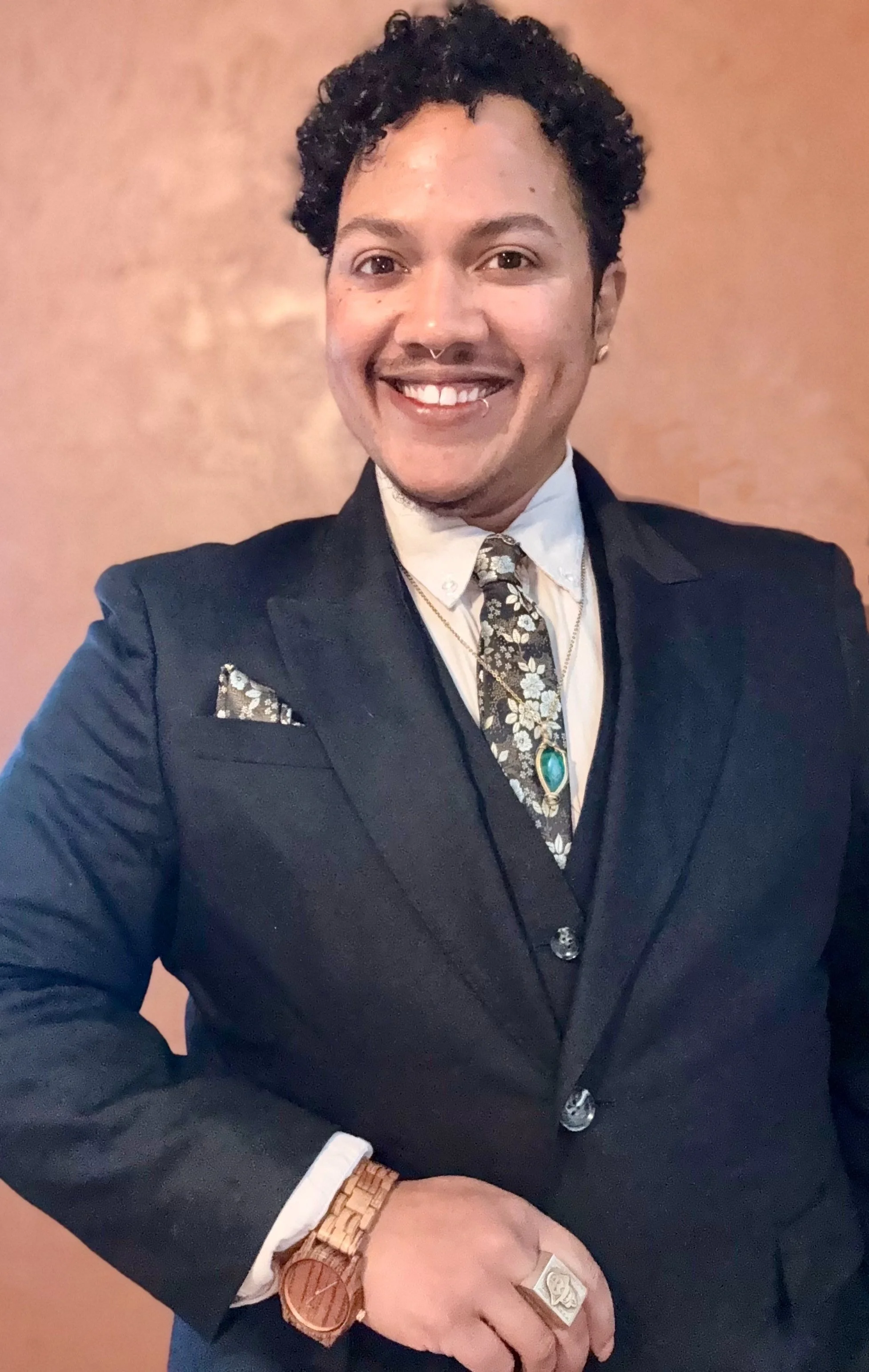 A Black trans man dressed in a formal black suit, white shirt, floral tie, and accessories including a turquoise pendant, ring, watch, and pocket square, standing against a plain background.