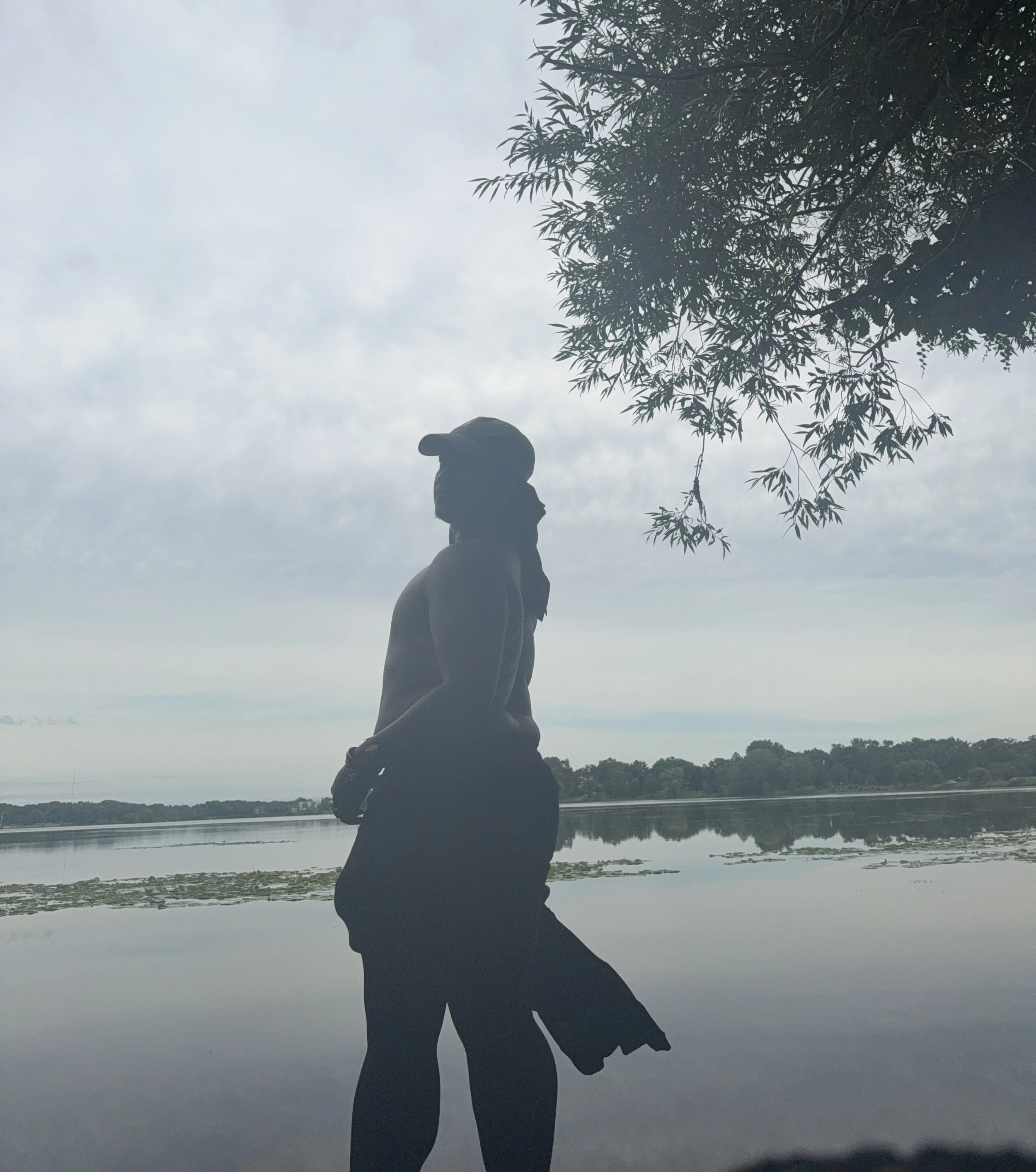 Silhouette of a Black trans man standing by a lake with water lilies, under a cloudy sky and tree branches overhead.