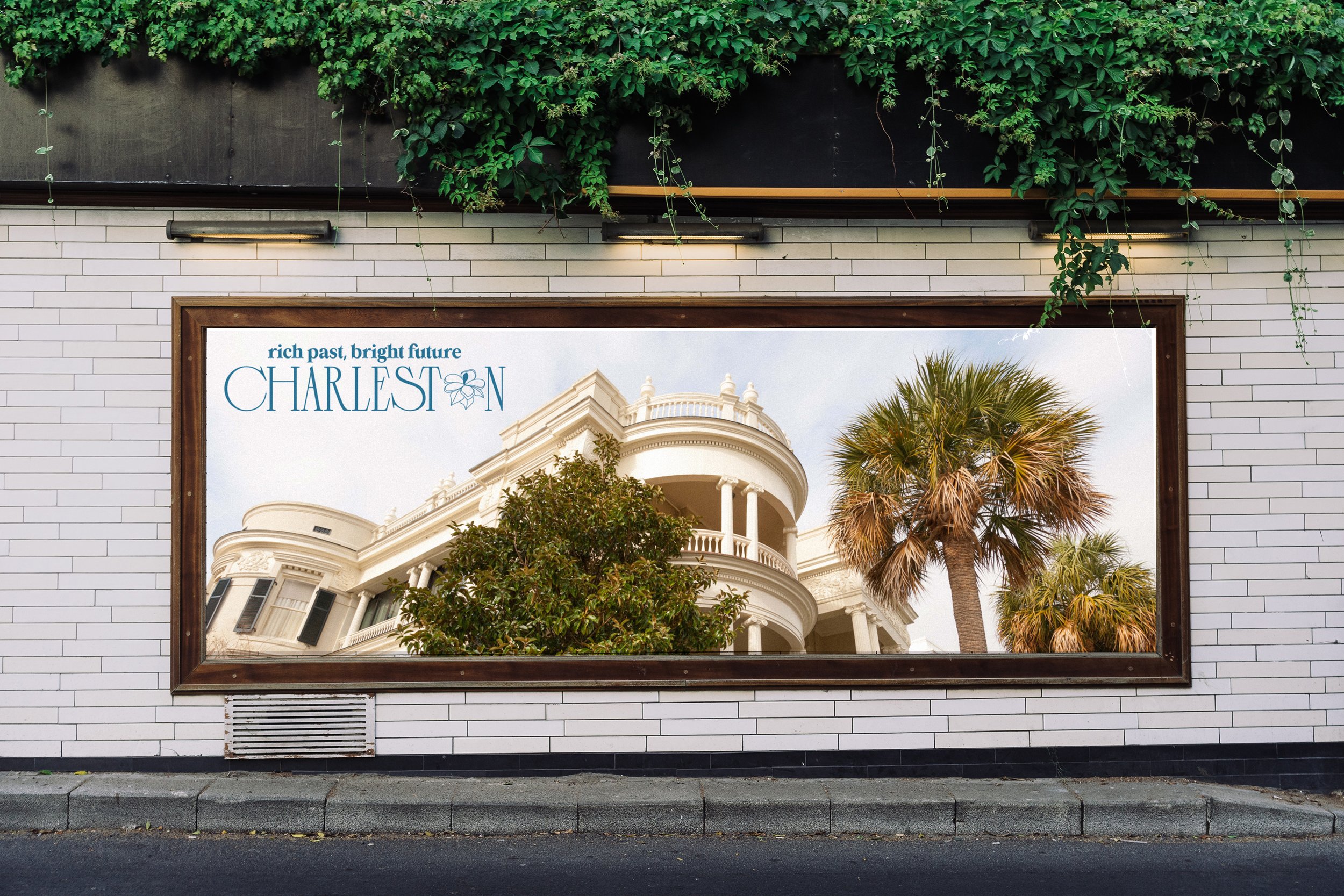 An outdoor white tiled wall on a city street that features a billboard and green vines hanging above it. The Charleston branded billboard features the Charleston logo and a photo of architecture and trees found there.