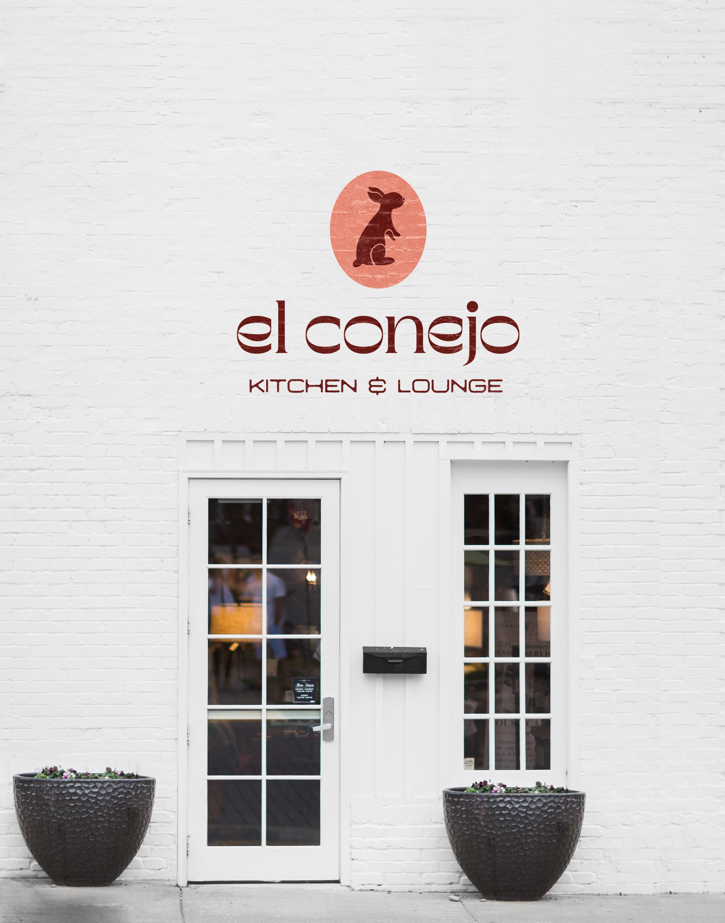 The El Conejo Logo painted on the exterior white brick front wall of the restaurant. Two planter pots are shown in front of two glass-paned doors.