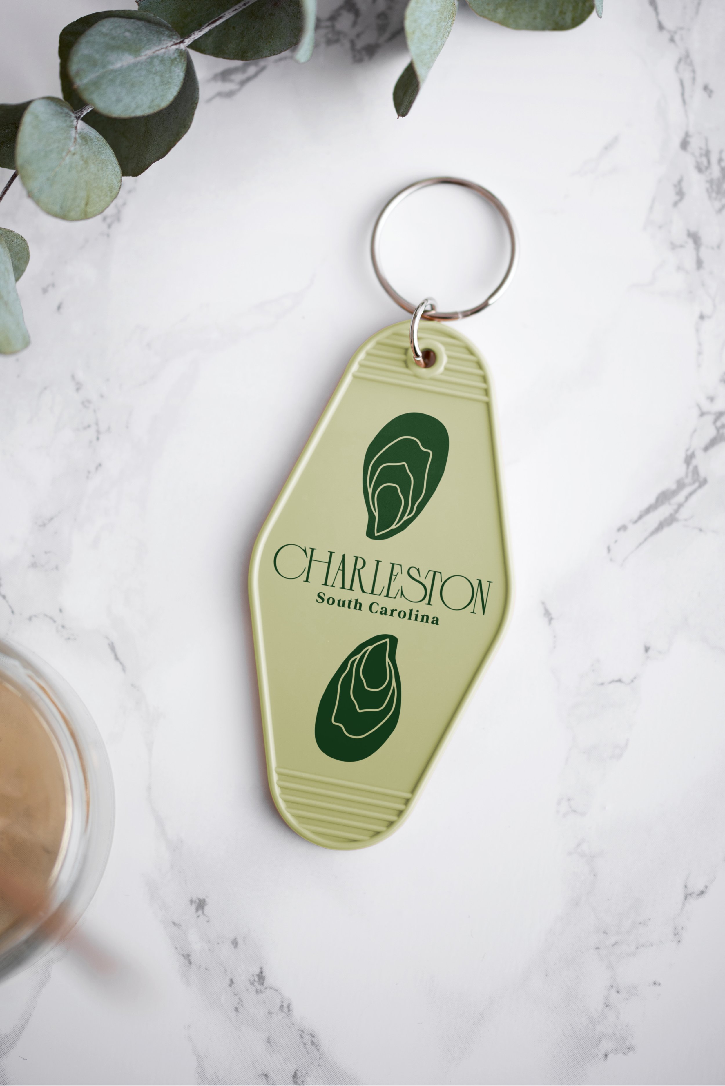 A close up photo of a light green Charleston branded keychain that has the Charleston logo above the words "South Carolina" and features two illustrations of oysters.