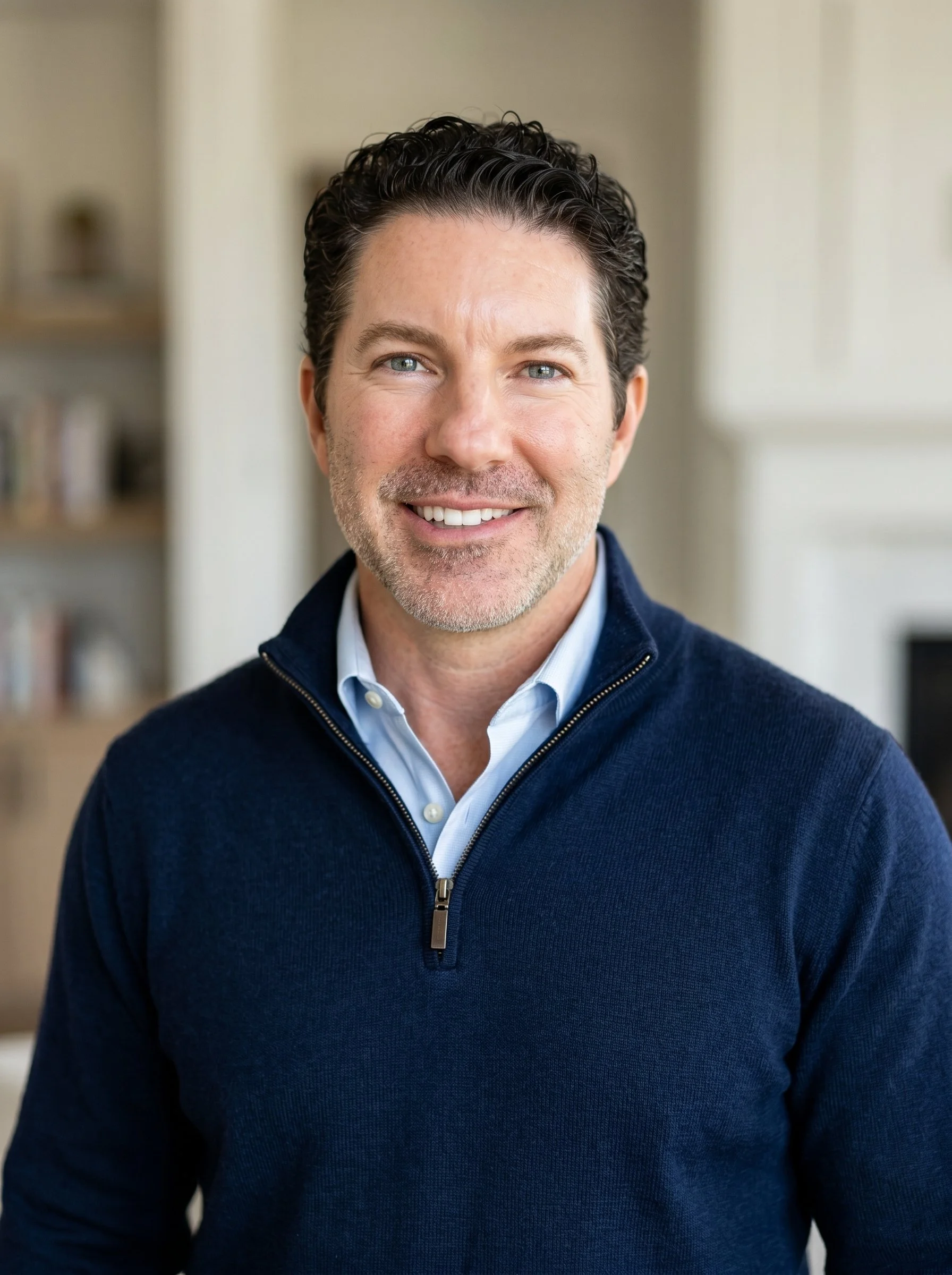 Close-up portrait of a smiling man with wavy dark hair, blue eyes, wearing a navy blue zip-up sweater over a light blue collared shirt, indoors with blurred background.