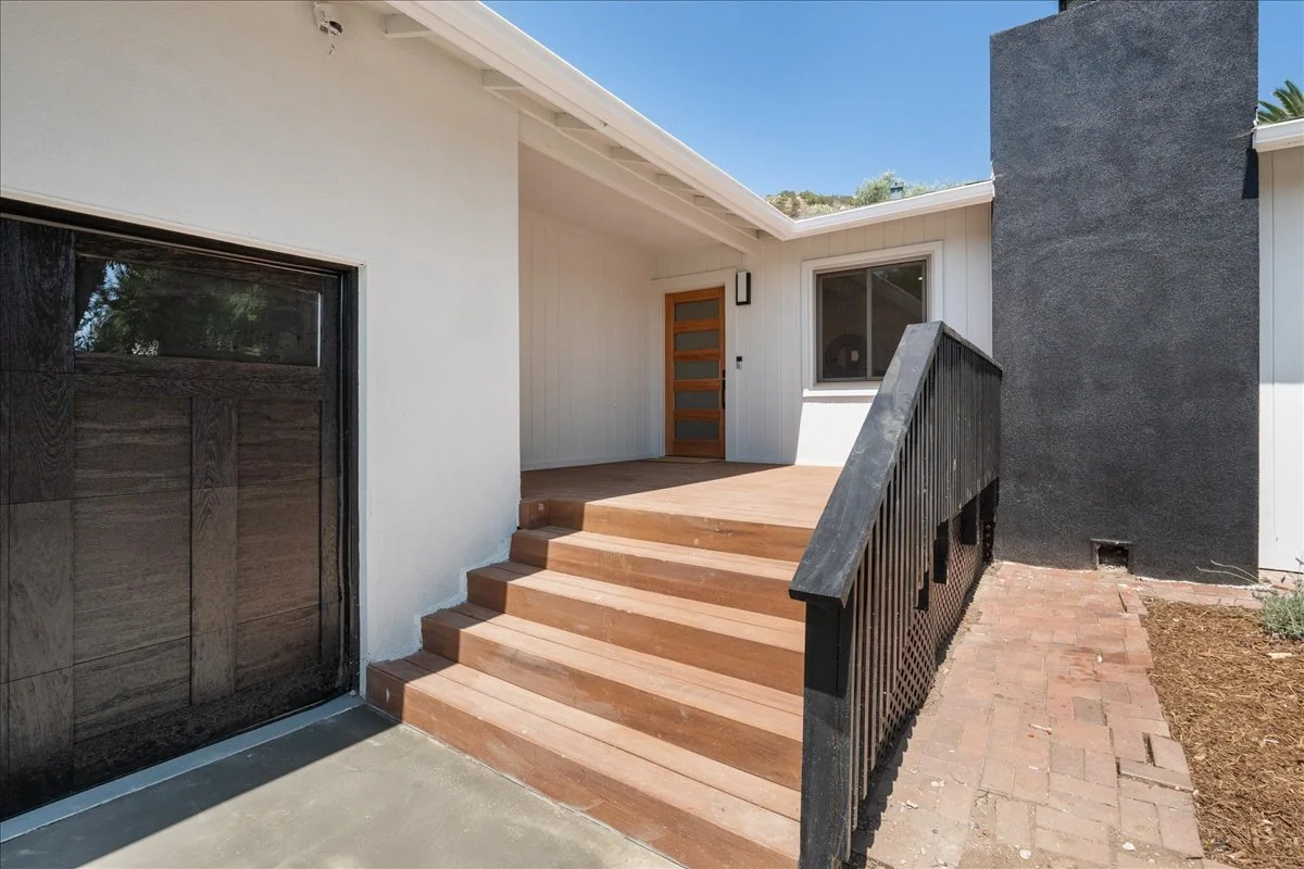 Front entrance of a modern house with wooden steps leading to the door, a black garage door to the left, white siding, a single window, and a black wall on the right, with a small garden bed and brick pathway outside.