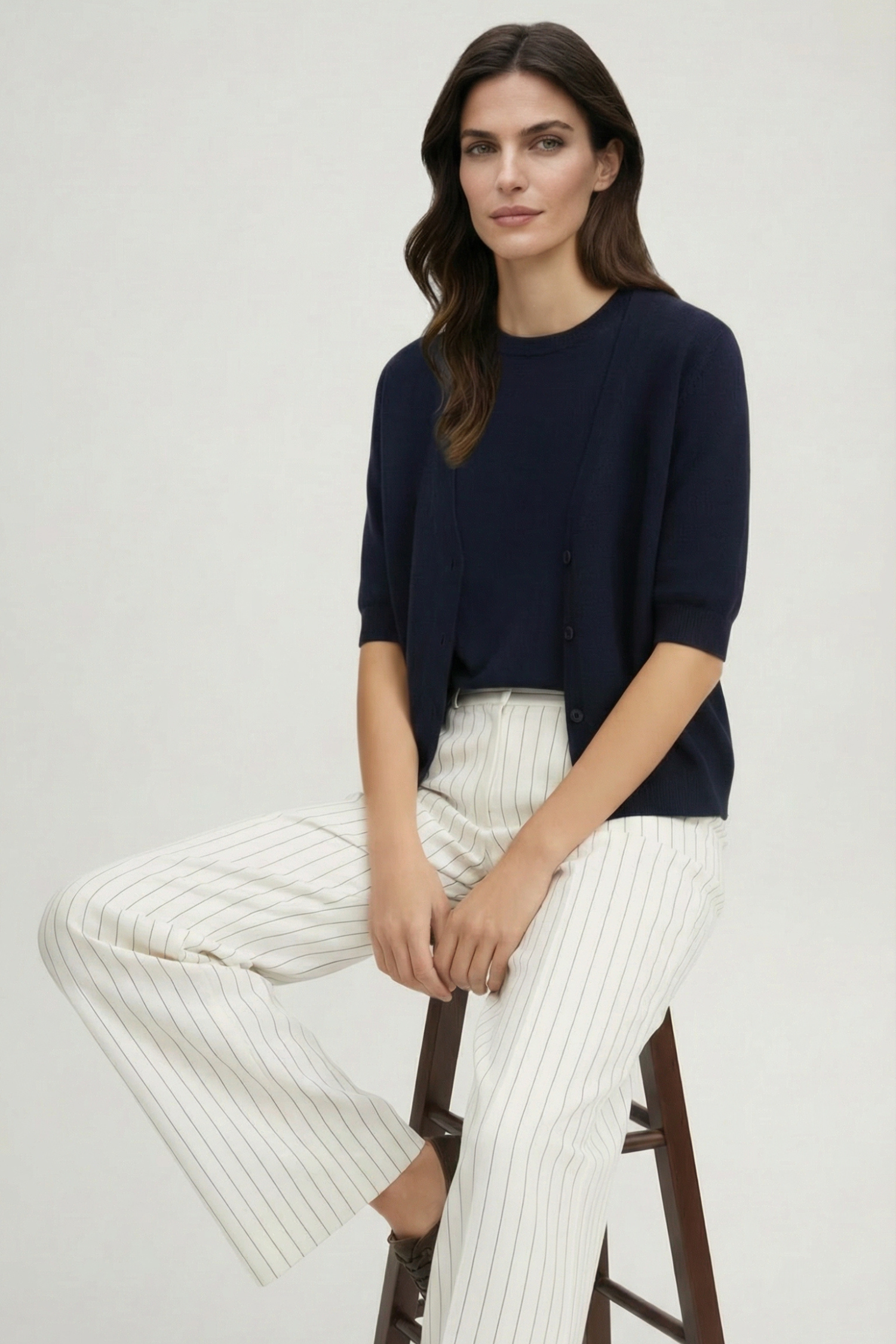 Brunette model sitting on a stool in a photography studio wearing a short sleeve navy cardigan set and white with navy pinstripe wide leg pants