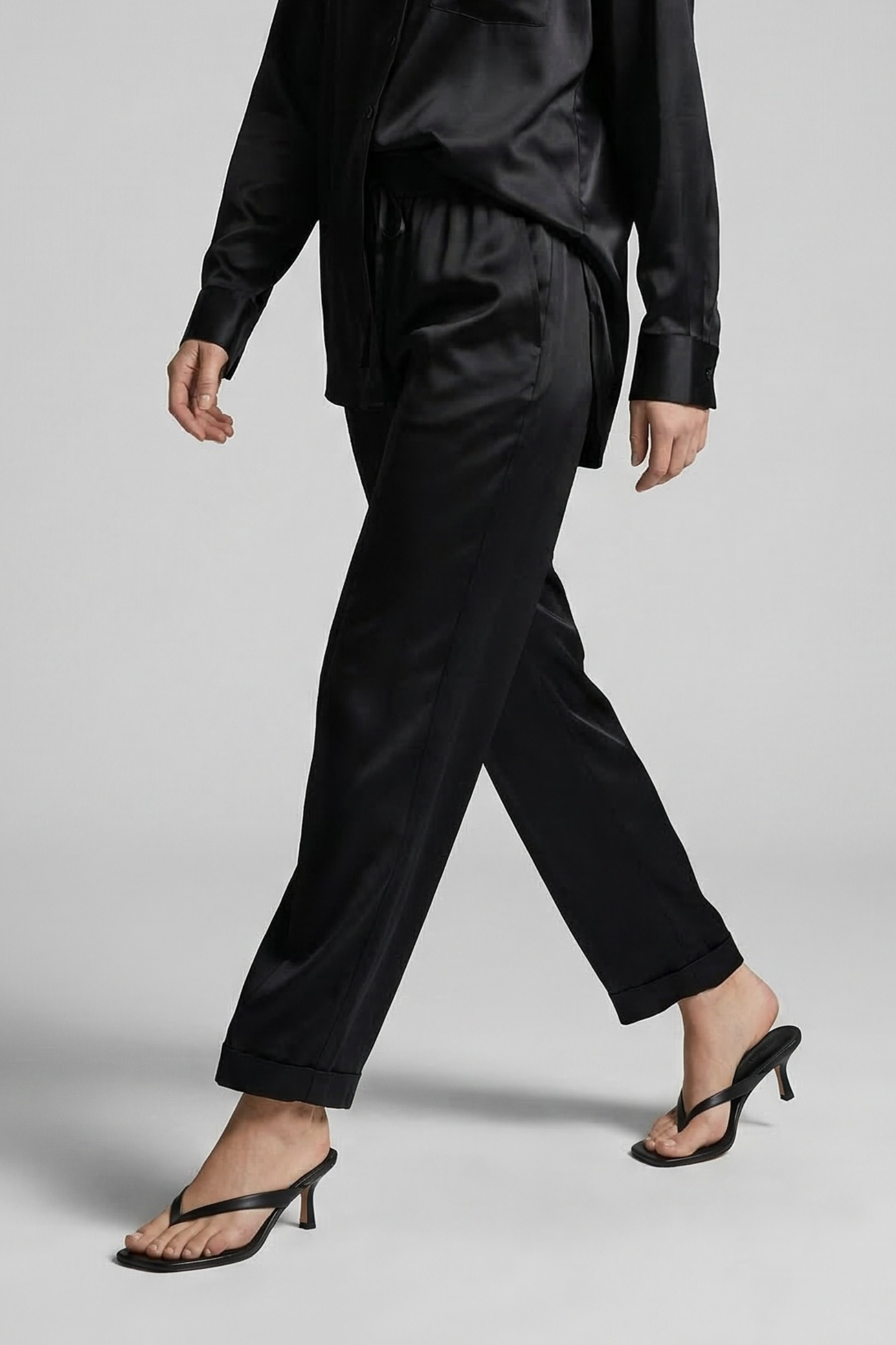 Model walking through a studio setting wearing a black silk lounge set and black heeled thong sandals