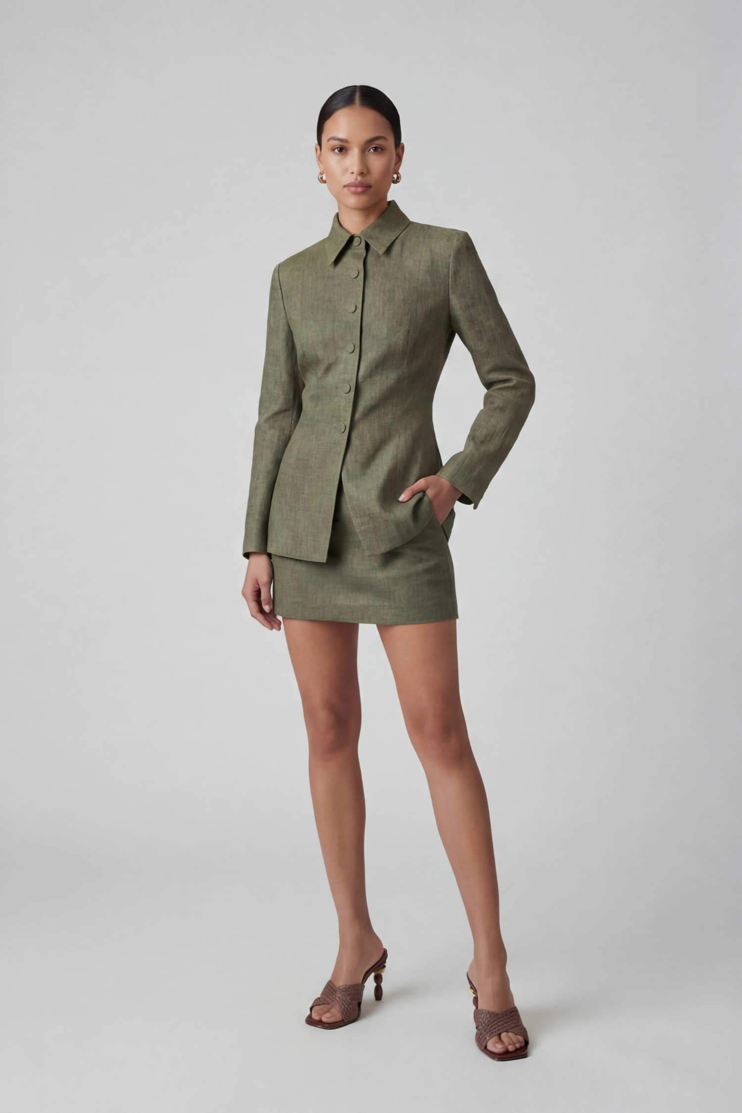 Brunette model with her hair pulled back standing in a photography studio wearing a green linen jacket and green linen mini skirt