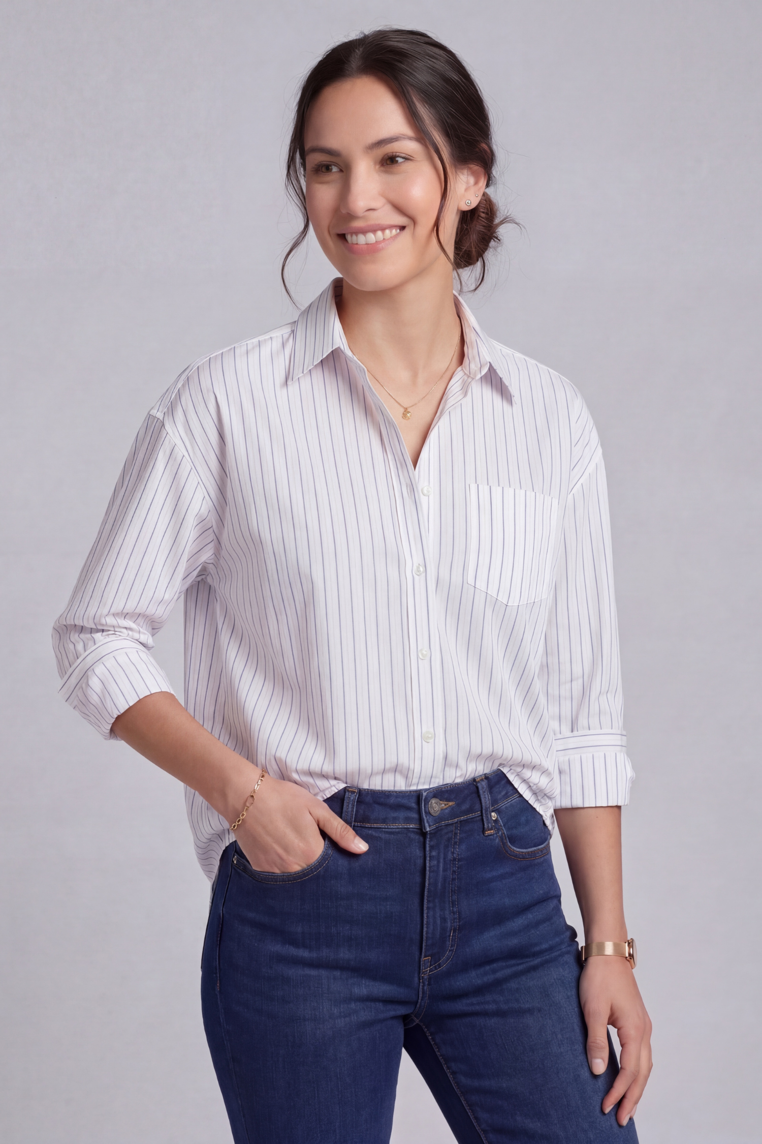 Brunette model with her hair in a low bun standing in a photography studio wearing a slightly oversized white and blue pencil stripe shirt and dark denim jeans