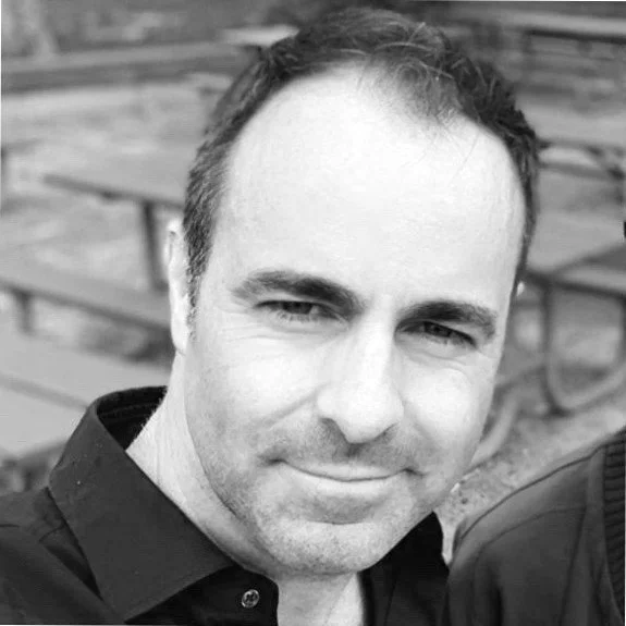 Black and white photo of a man with short, dark hair, wearing a collared shirt, smiling slightly, outdoors with a picnic table in the background.