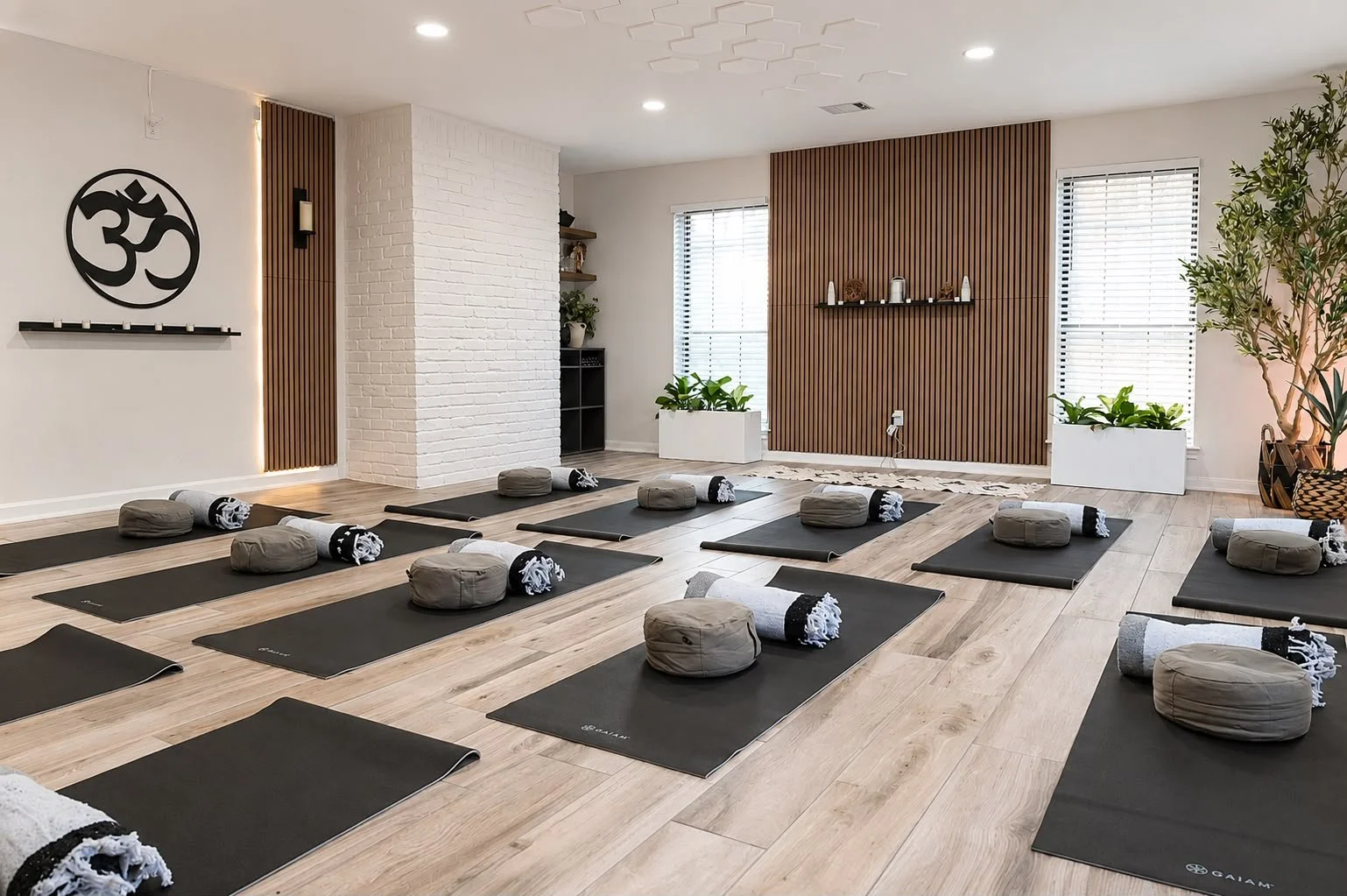 Yoga class in a studio with black mats, bolsters, and blankets, decorated with plants, wood panel accents, and a white brick wall with a black Om symbol.