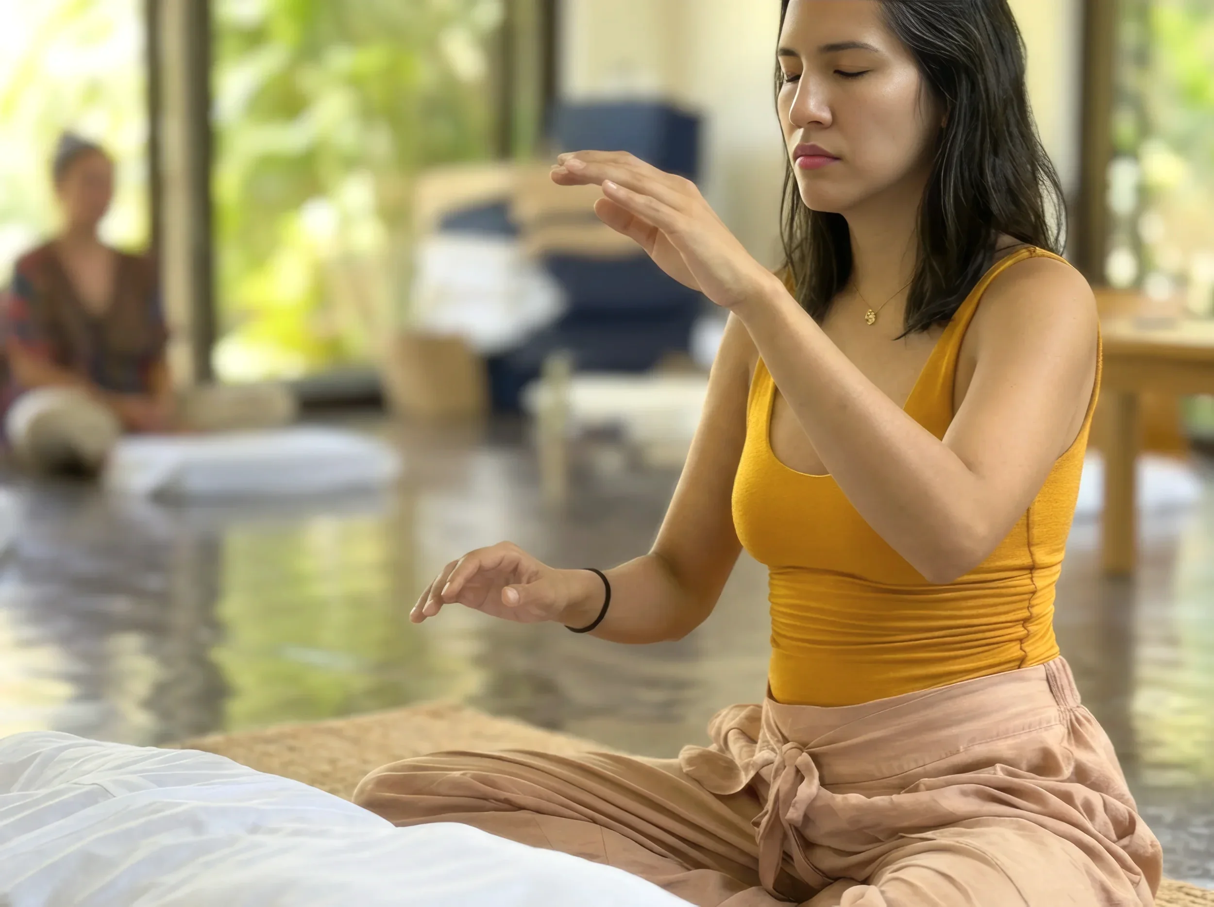 A woman in a yellow top practicing yoga or meditation indoors near large windows with greenery outside.