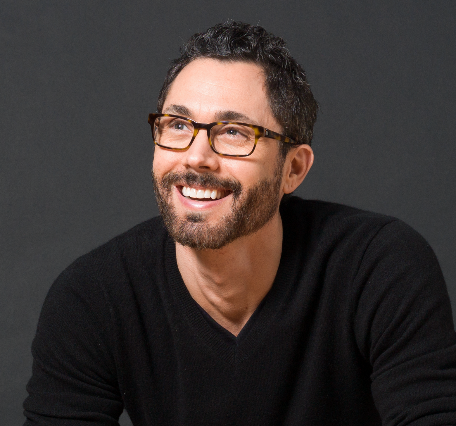 A smiling man with dark, curly hair and glasses wearing a black shirt, posing against a gray background.