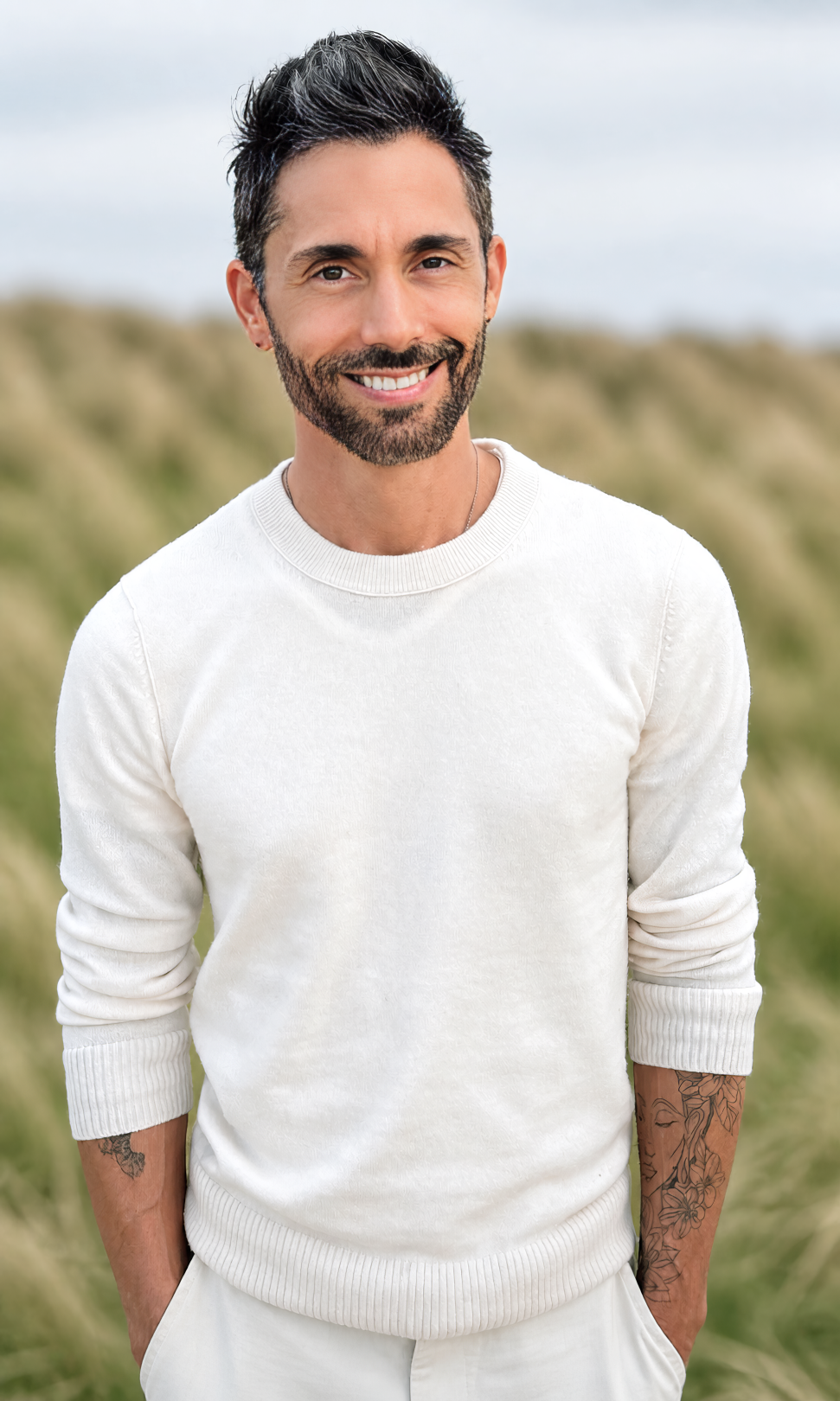 A smiling man with dark hair, a beard, and tattoos on his arms, wearing a white sweater, standing outdoors in a grassy area with a cloudy sky.