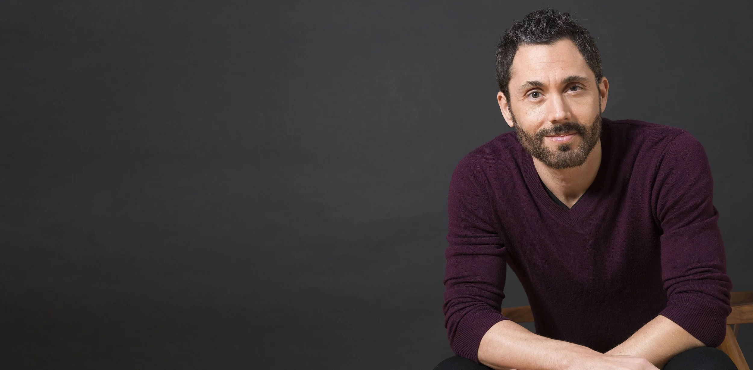 A man with dark hair and a beard wearing a maroon sweater, sitting against a dark background.
