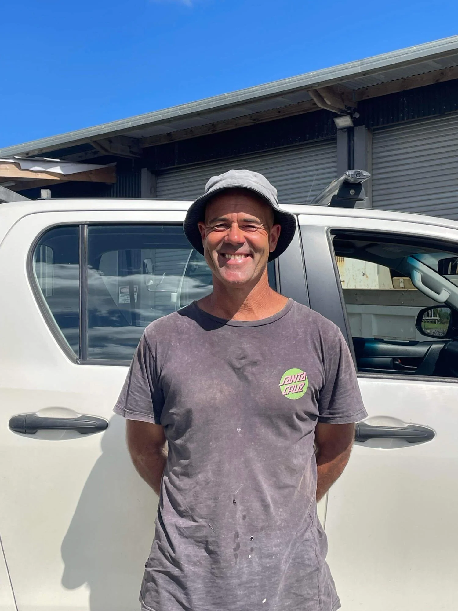 Joe O'Malley smiling in front of a white vehicle with a building in the background, wearing a hat and a dark t-shirt.
