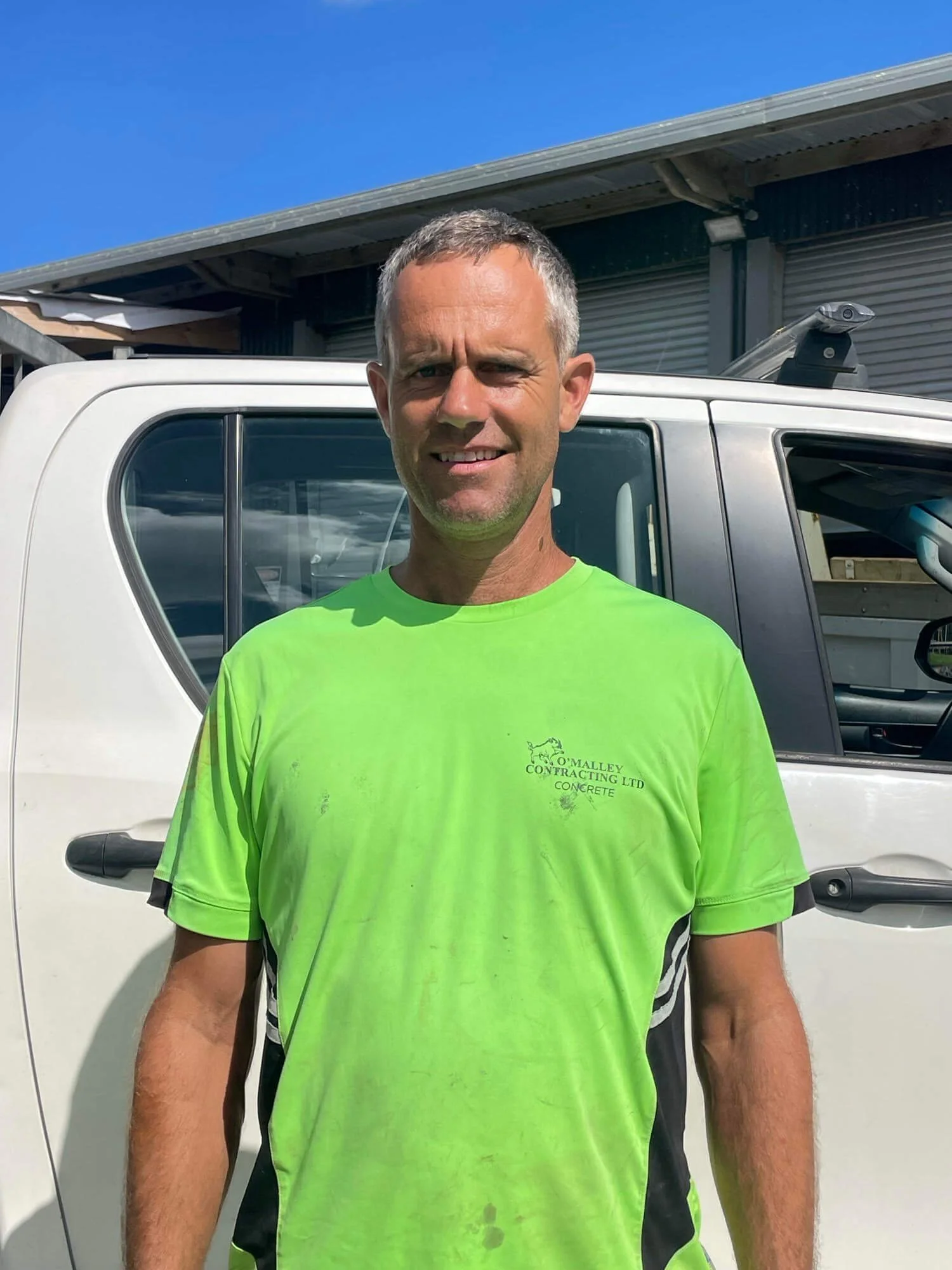 Kevin O'Malley standing in front of a white vehicle, wearing a bright green T-shirt with a company logo, outdoor setting in Kaiwaka.