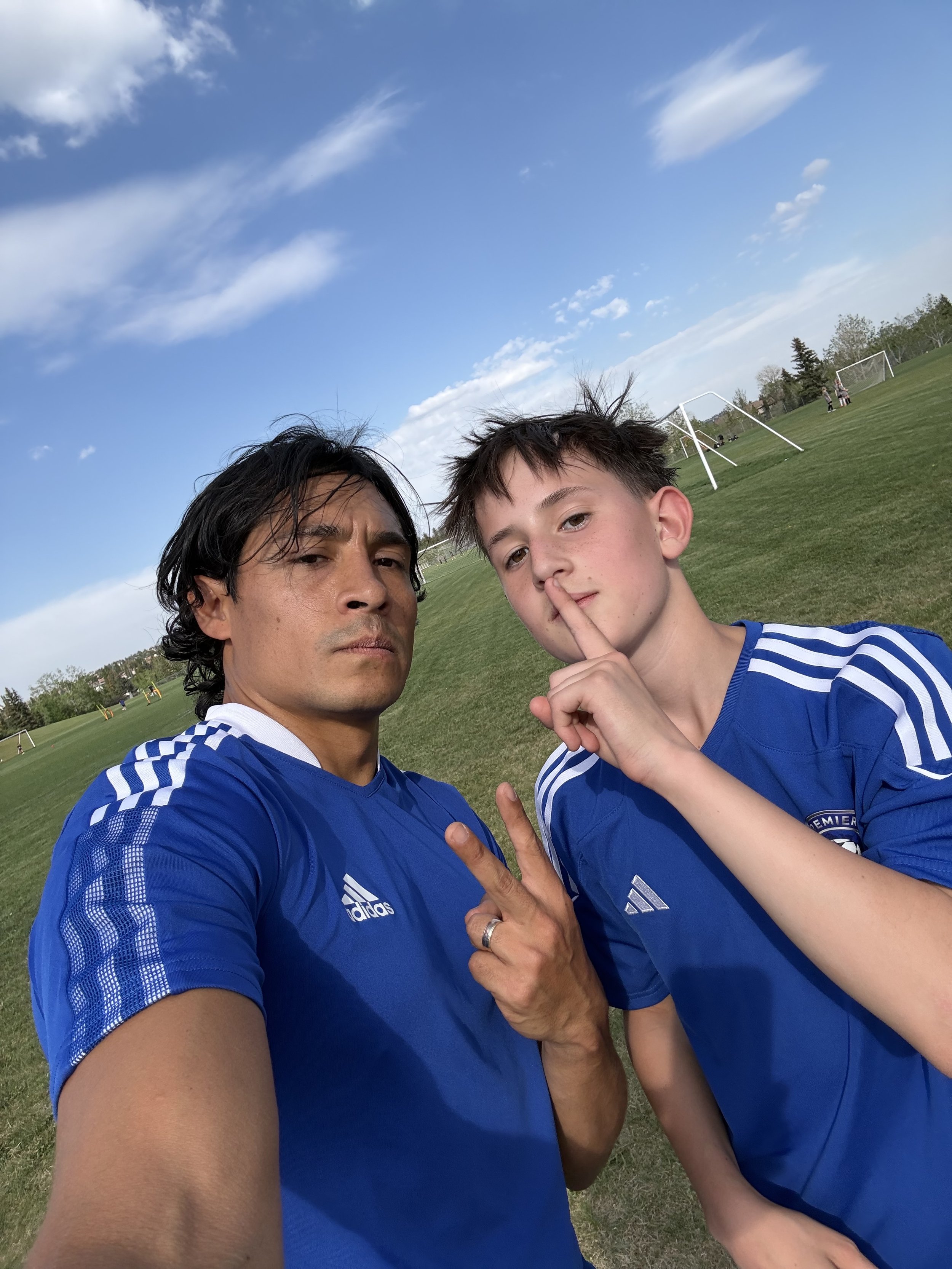 Two males taking a selfie in an indoor sports court; one man with dark hair, wearing a black shirt, making a peace sign, and a young boy with short brown hair, also in a black shirt, smiling.