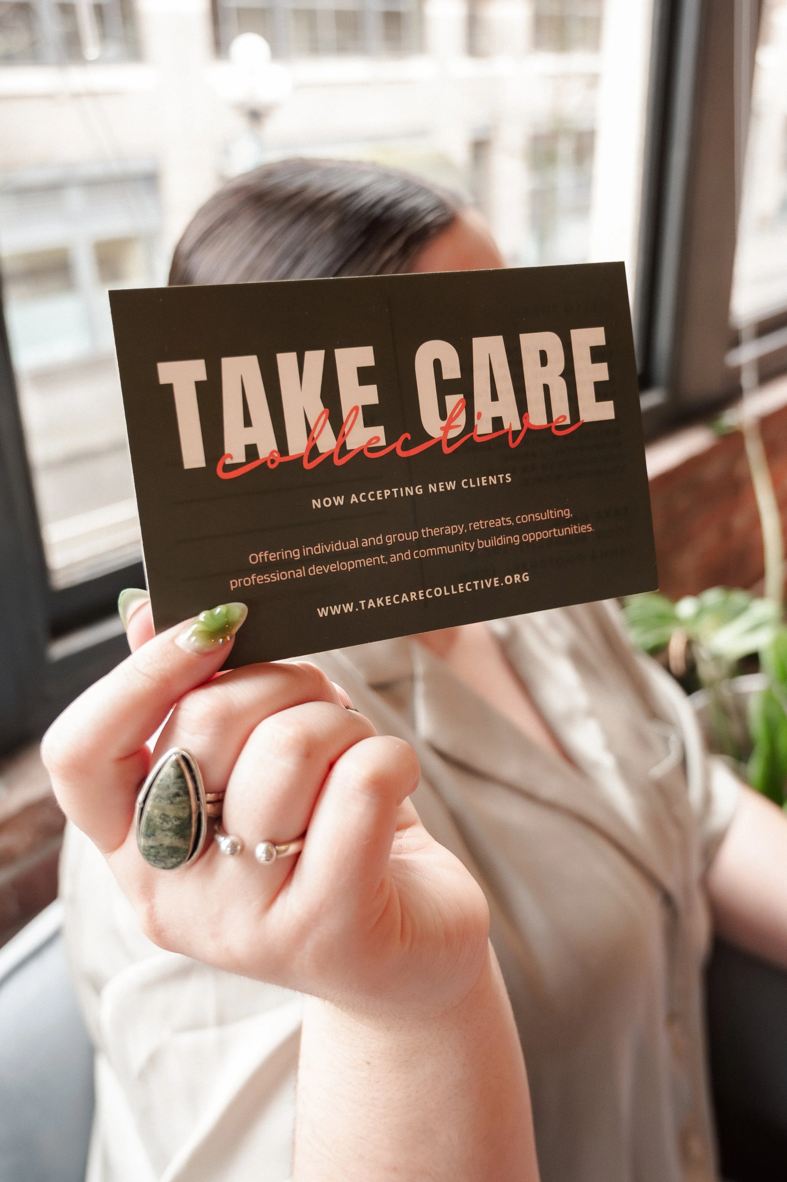 Person holding a black card with white and red text that reads ‘Take Care Collective,’ with additional information about accepting new clients and a website URL, partially visible, in the background.