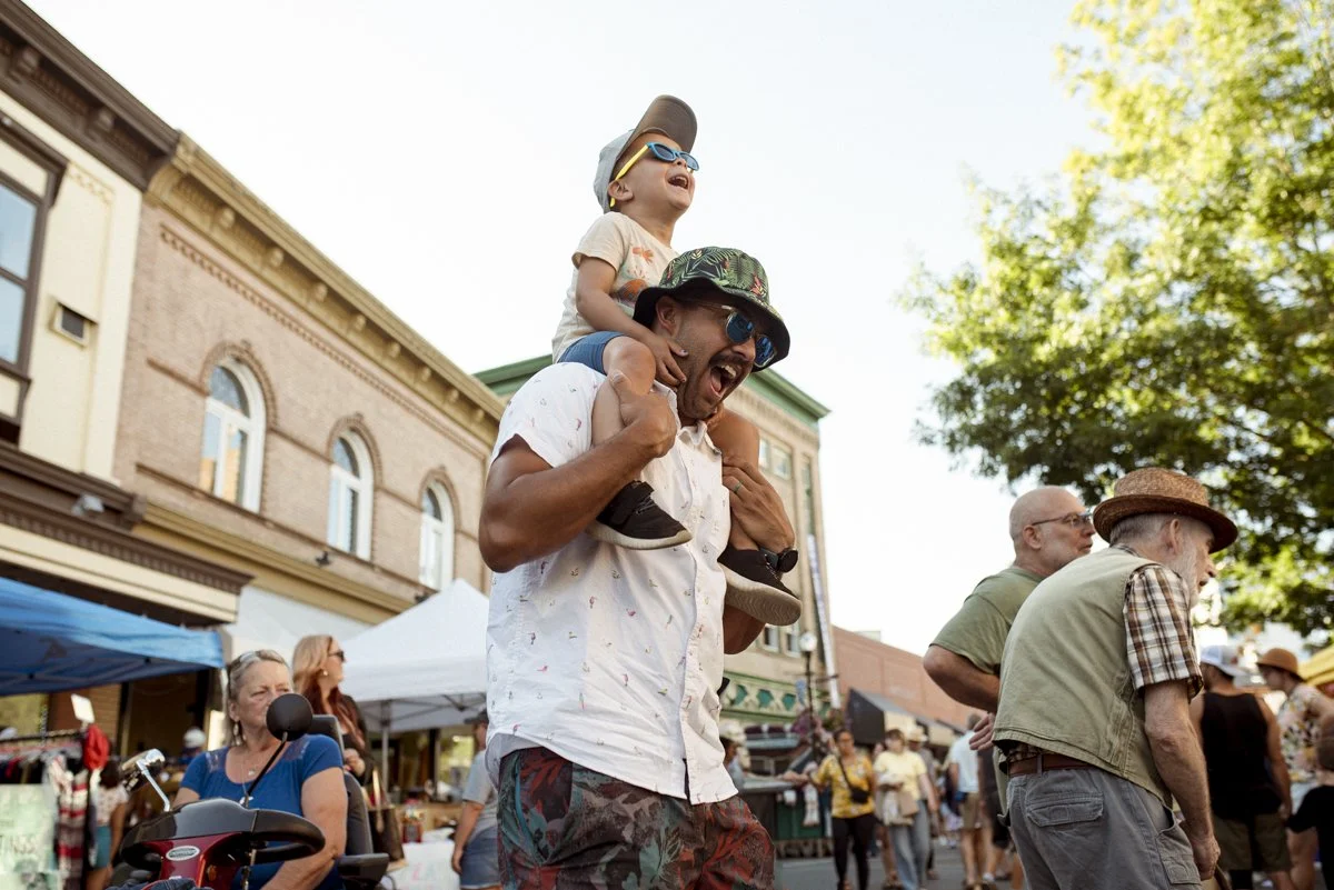 A father carrying his son on his shoulders at a street event. There are several people around them on the street, including a person using a mobility scooter and to men standing to the right of the image one of them is wearing a hat.