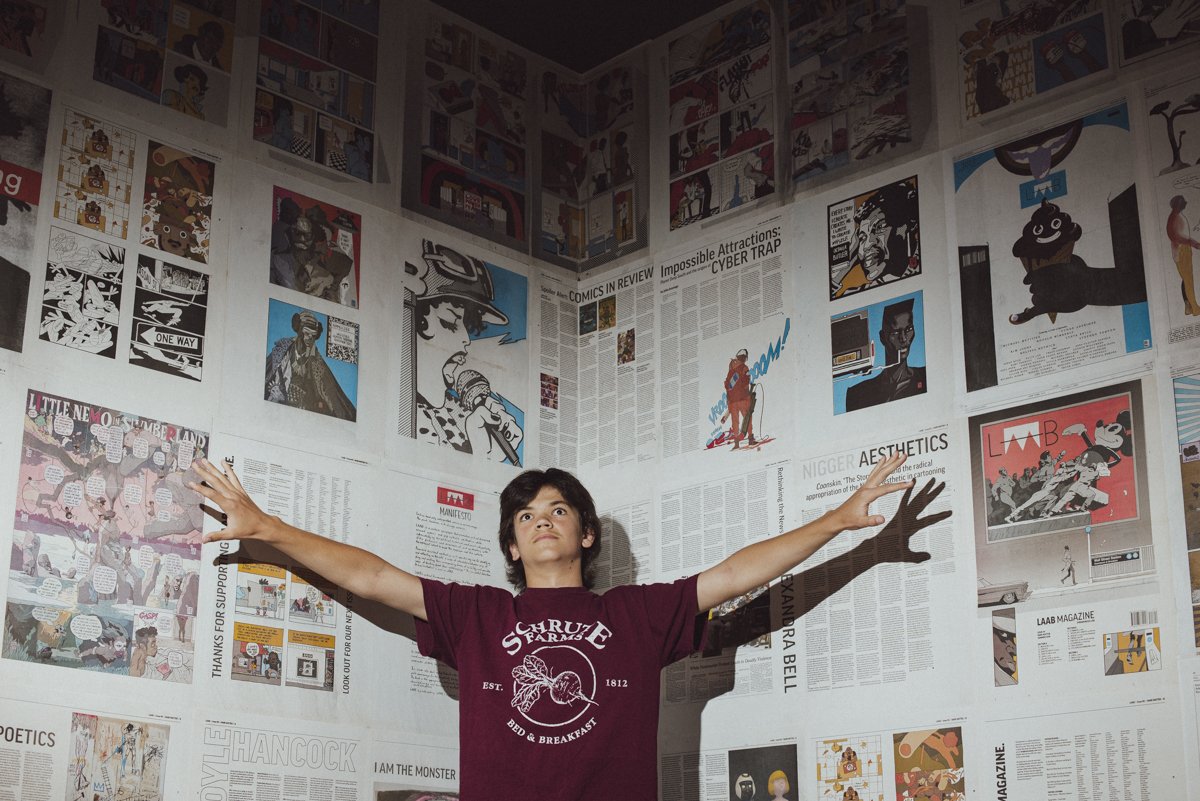 A young person standing in a museum with their arms stretched out wide. The background is a wall full of comic strips and newspapers.