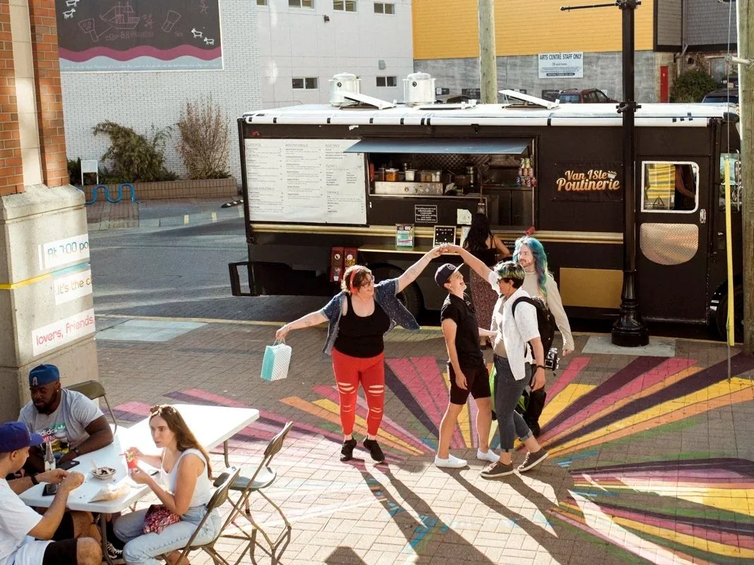 Friends dancing in the sunlight at an outdoor event. There is a food truck in the background and people sitting at a table enjoying food in the bottom right corner.