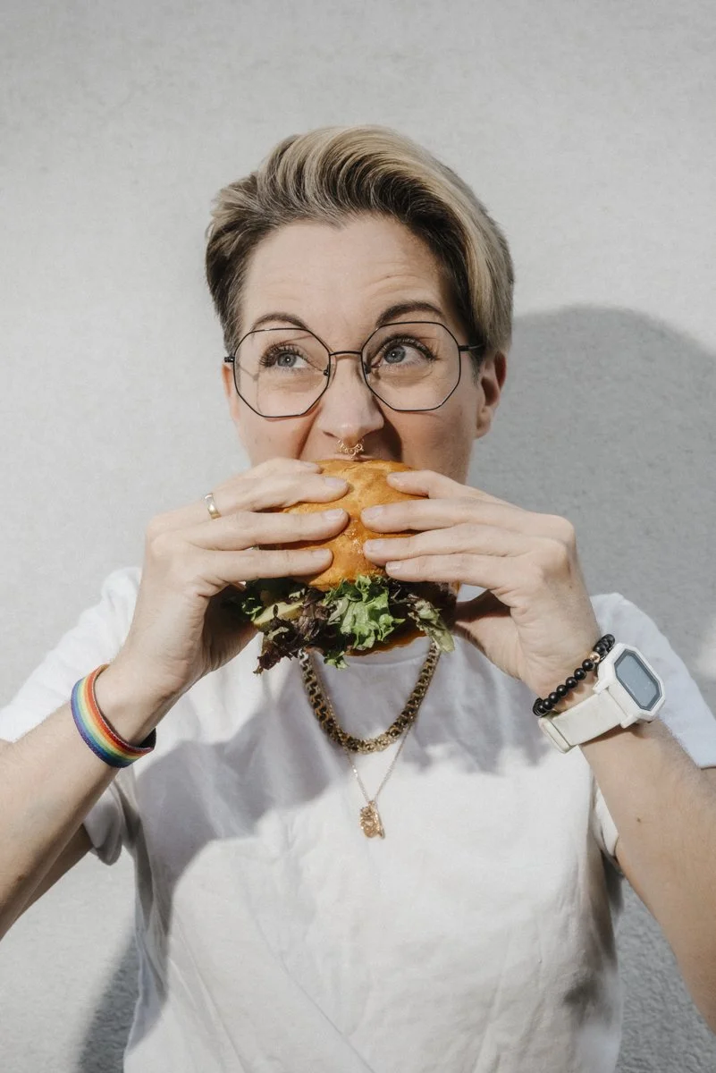 A person taking a big bite out of a hamburger. they have short hair, they are wearing glasses, they have a rainbow bracelet on one wrist and a black beaded bracelet and white watch on the other.