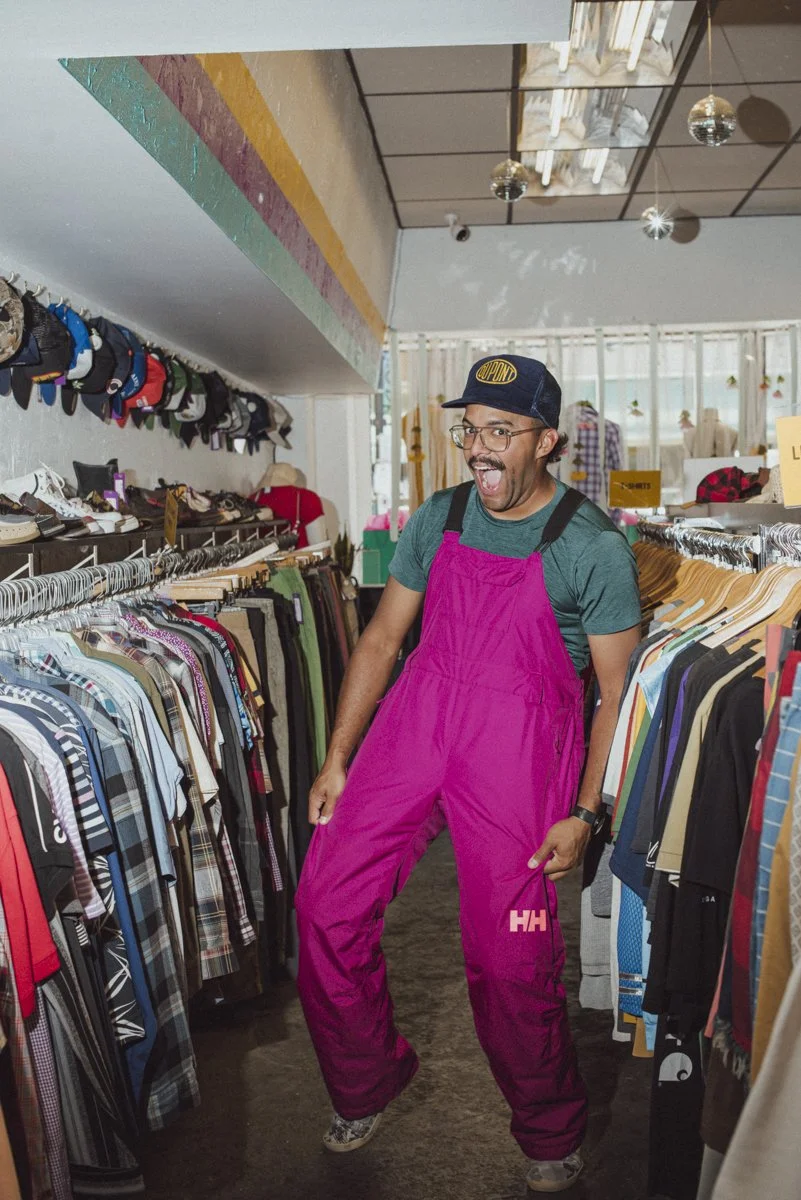 A person trying on pink ski pants in a consignment store. He is wearing a blue hat, glasses and is smiling. 