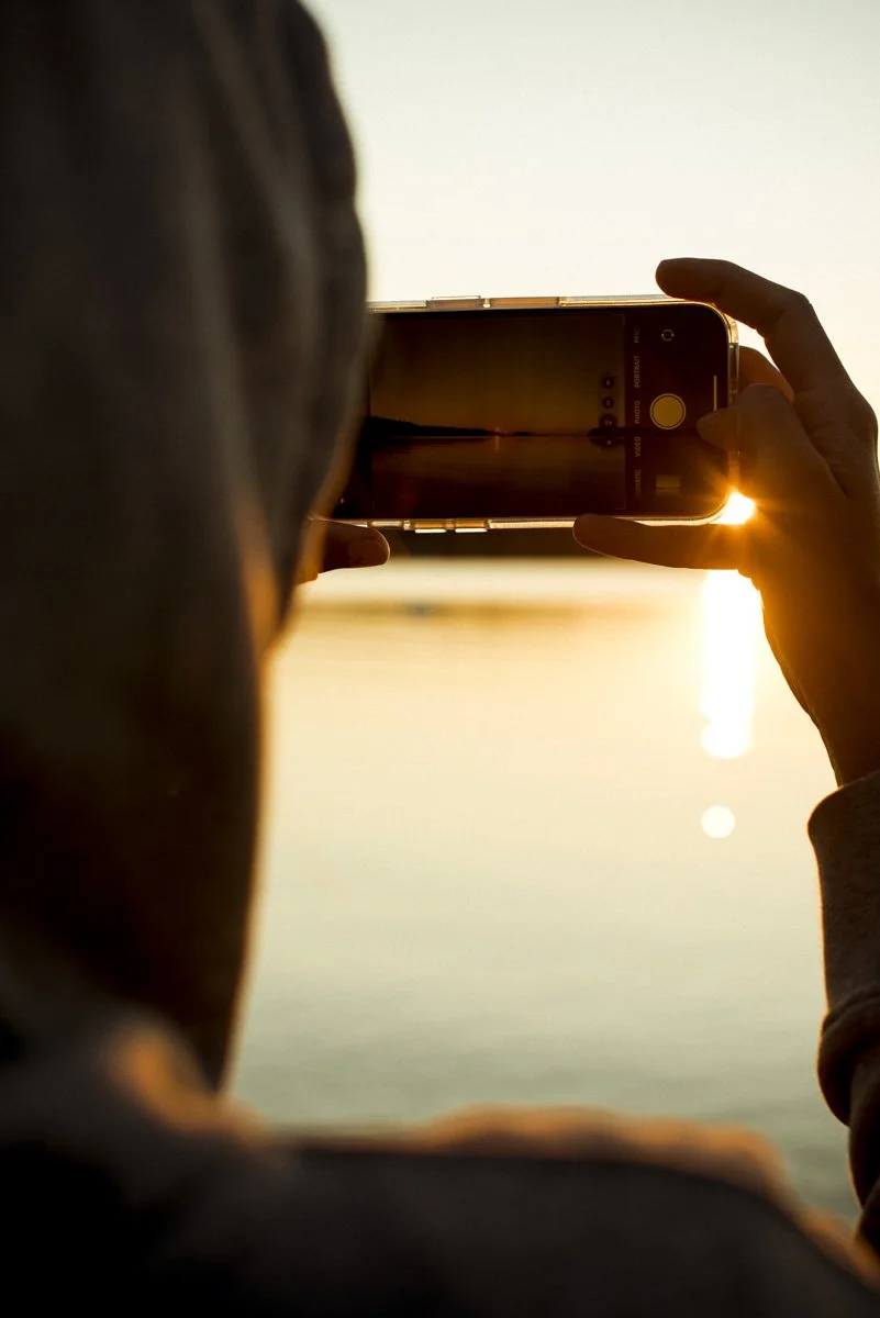 A person holding up their phone camera to watch the sun rise. The sun is shining through their fingers and reflecting on the water. 