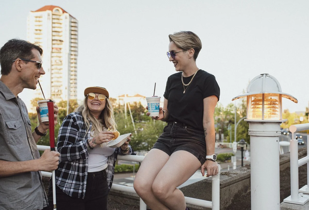 3 friends enjoying food and drinks, over looking the marina. One wearing yellow sunglasses and a hat, one wearing dark black sunglasses and carrying a mobility cane and the back of another friend who is wearing a black shirt and shorts.
