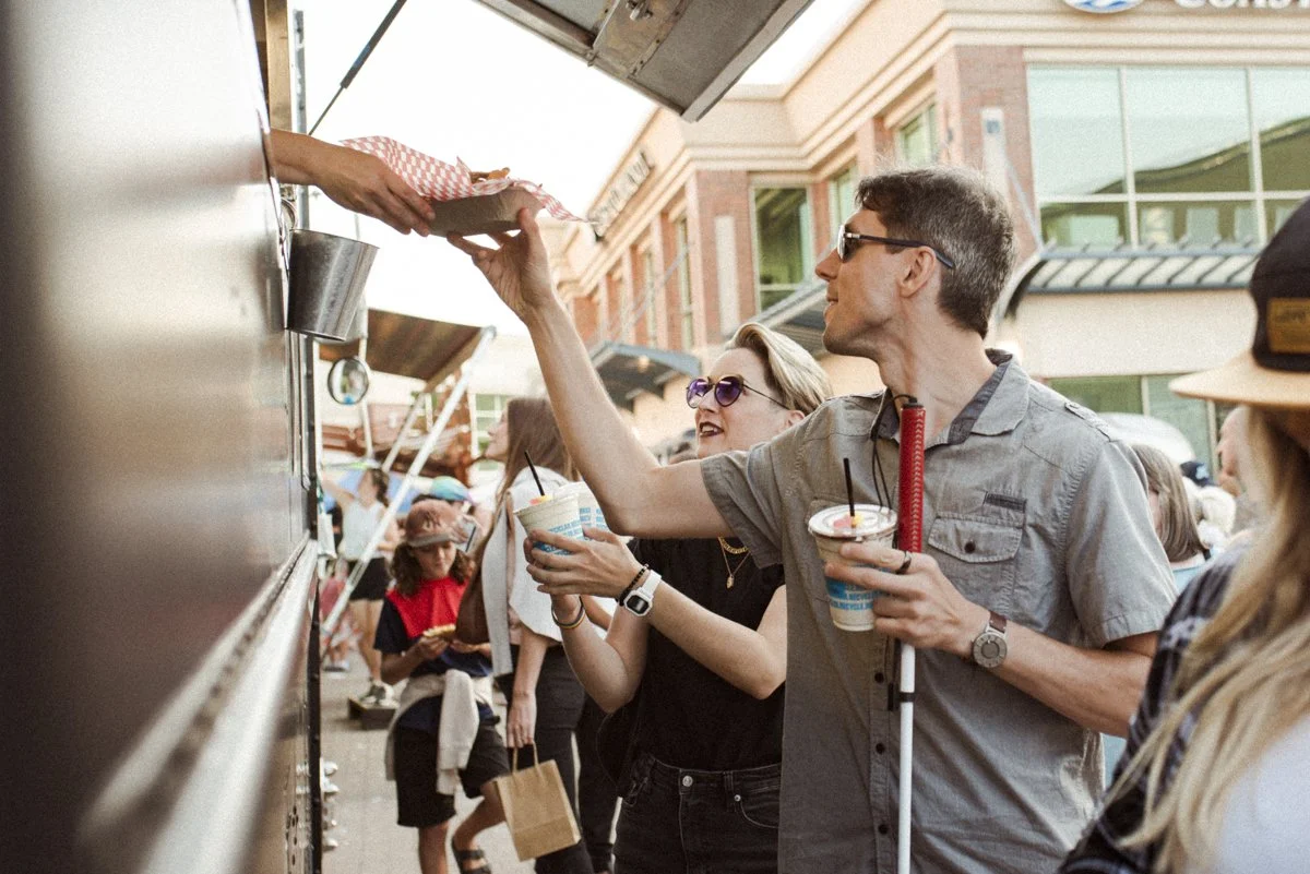 A man reaches for his food from a food truck with one hand. In his other hand he is holding a drink and his mobility cane.
