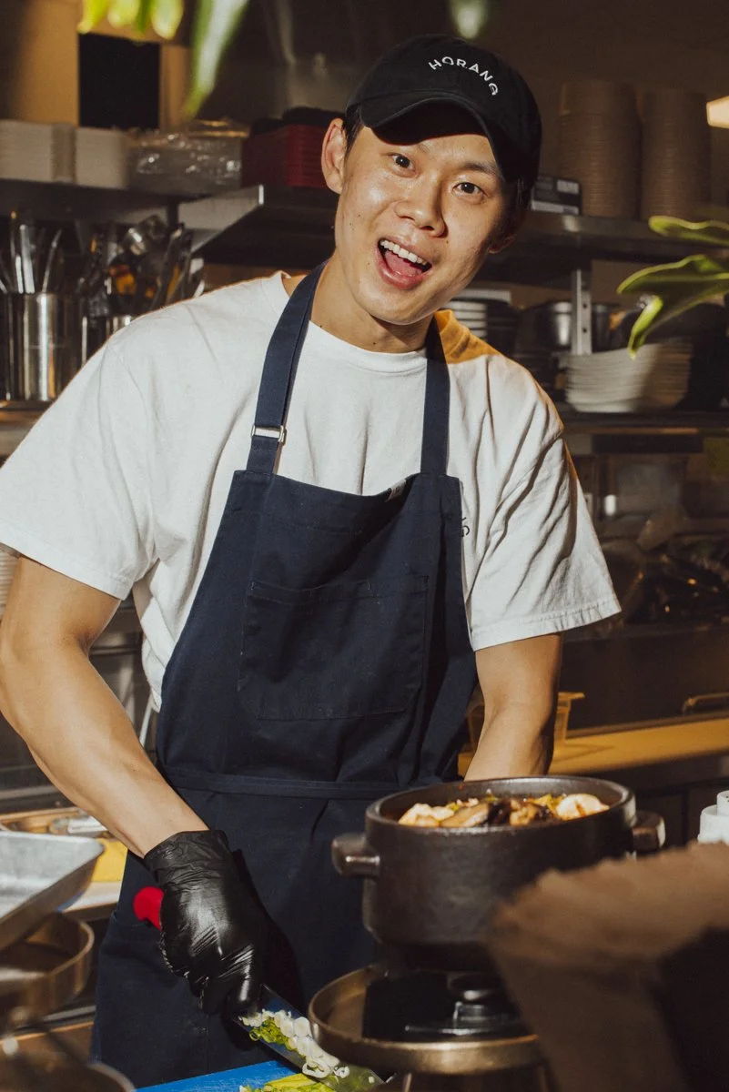 A chef making a meal wearing a black hat and black apron. in the background is the restaurant kitchen, in the foreground is a hot pot.