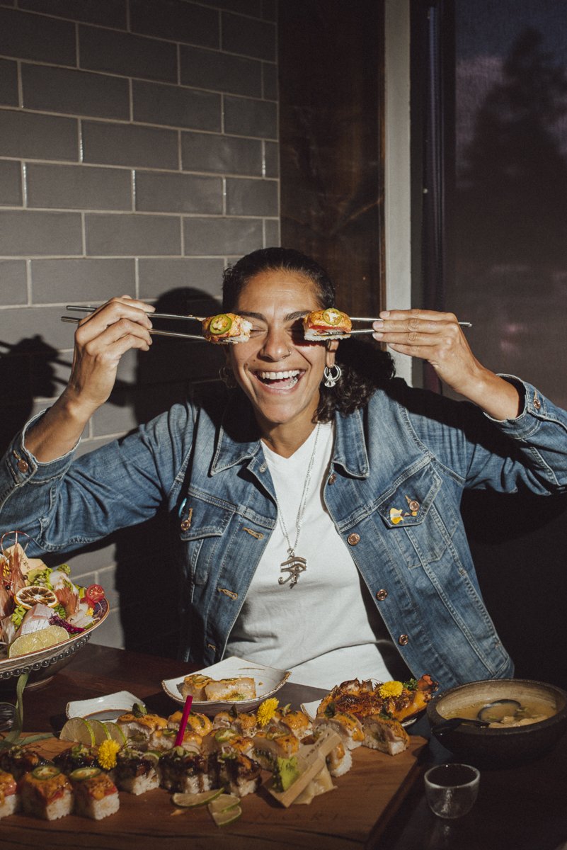 A woman eating sushi. She is holding two pieces of sushi up in front of her eyes. She is using chopsticks.