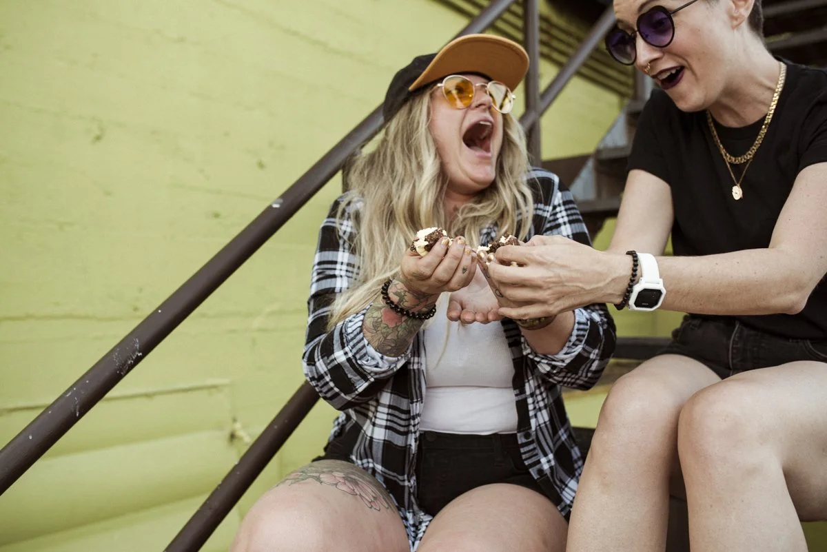 two friends laughing and sharing a dessert square on the back stairs of a green building.