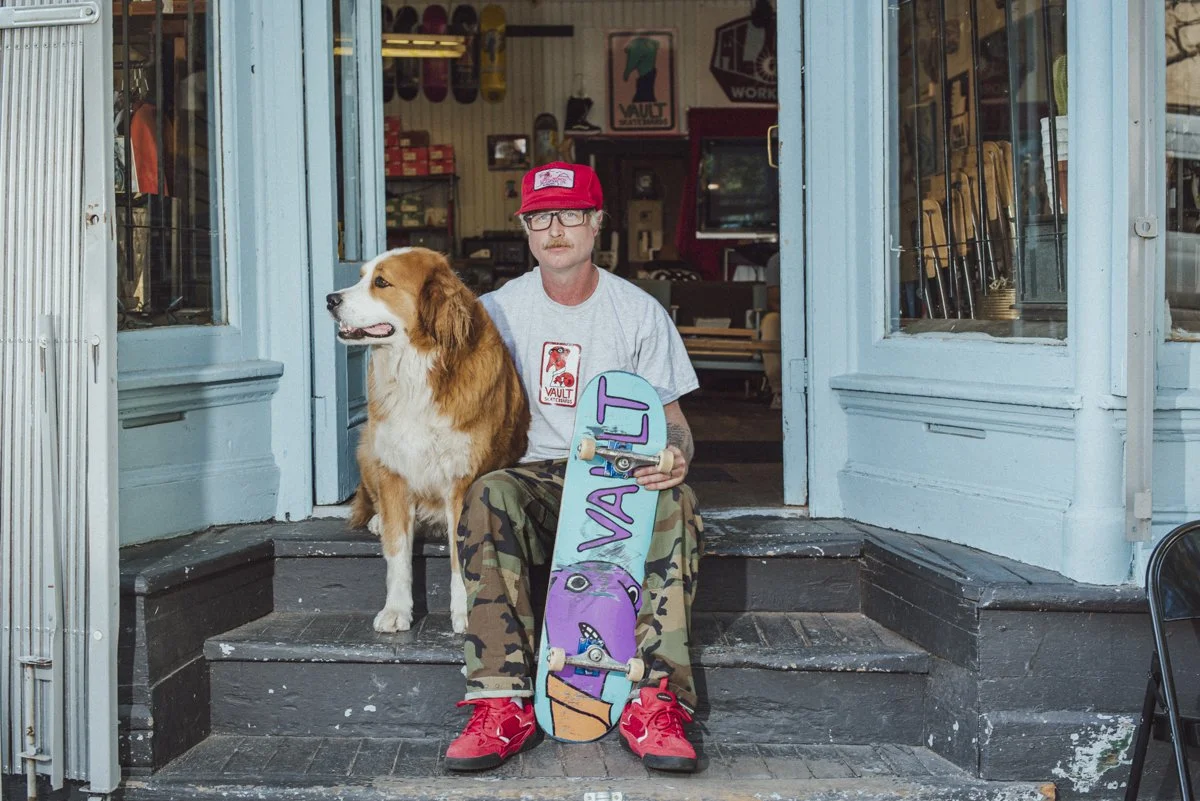 A shop owner and his dog sitting on the shop steps. The owner is wearing a red hat, has one hand around is dog and with the other hand he is holding a skateboard.