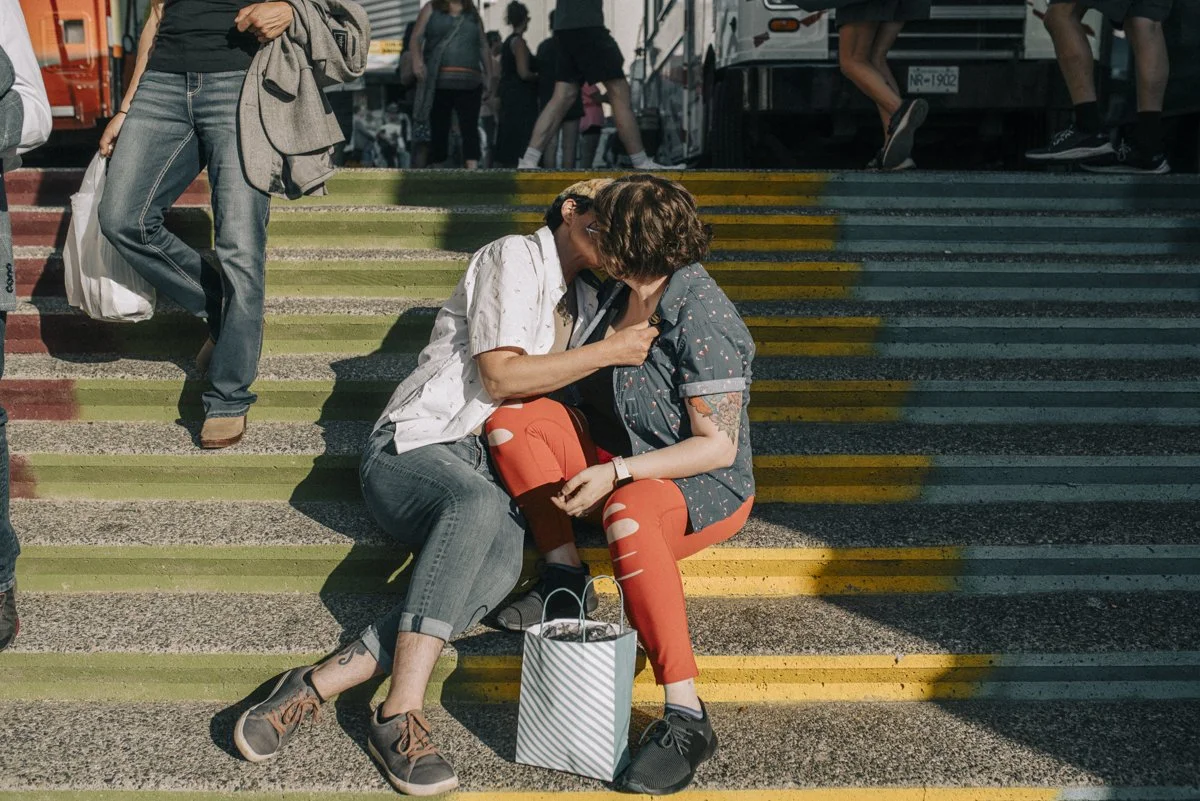 Two people kissing on rainbow steps as others walk past them.