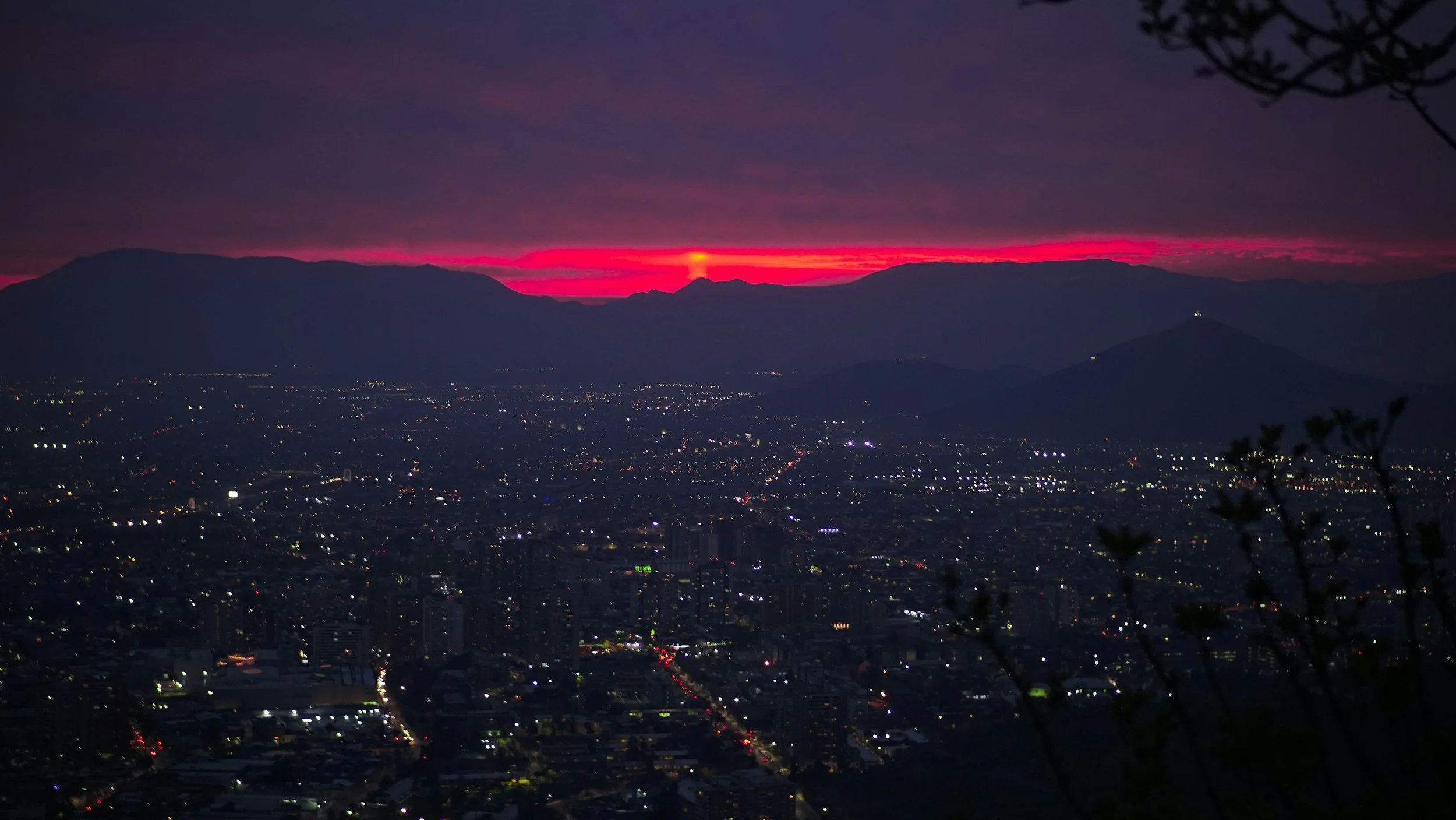 Cityscape at dusk with sunset sky, mountain silhouette in the background, and city lights in the foreground