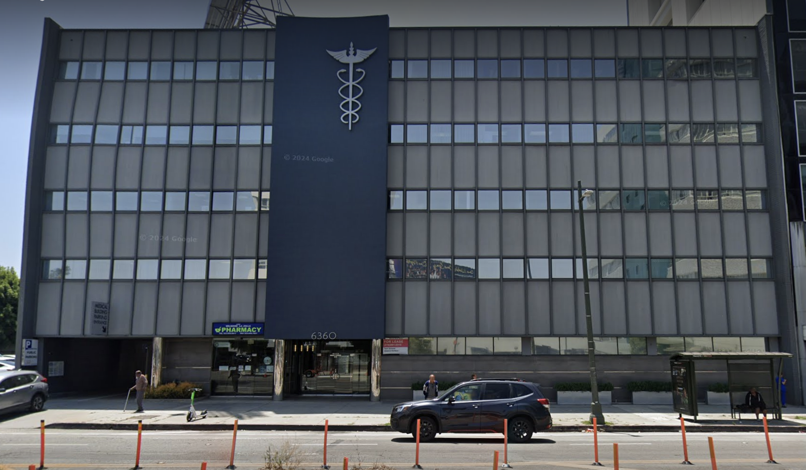 Front view of a multi-story office building with a healthcare symbol at the top, large windows, a pharmacy sign at the entrance, parked cars, and people walking on the sidewalk.