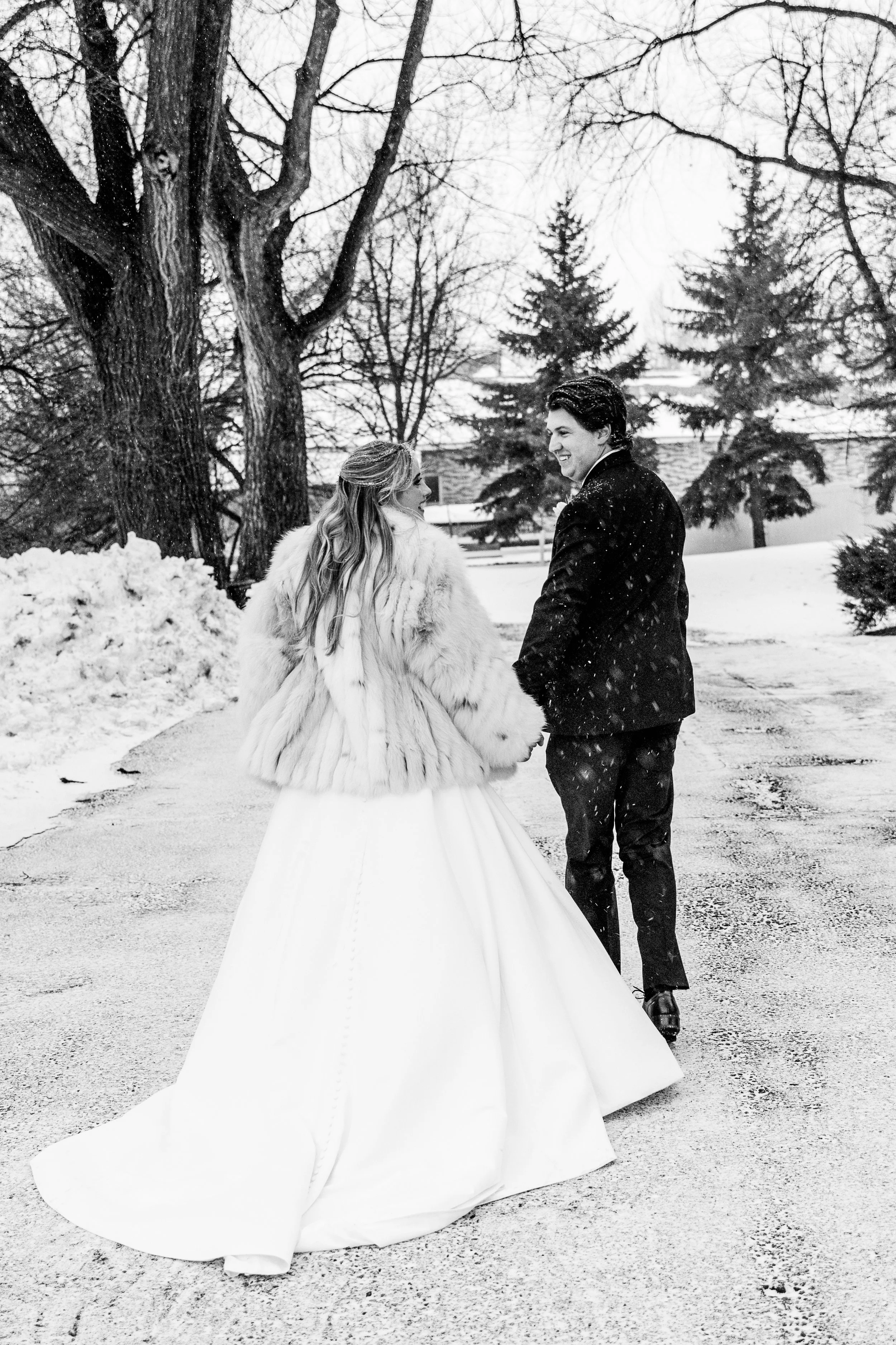 A newlywed couple holding hands and walking outdoors in a winter setting, with snow on the ground and leafless trees in the background. The bride is wearing a large white gown and fur coat, and the groom is dressed in a dark suit. Snow is falling aro