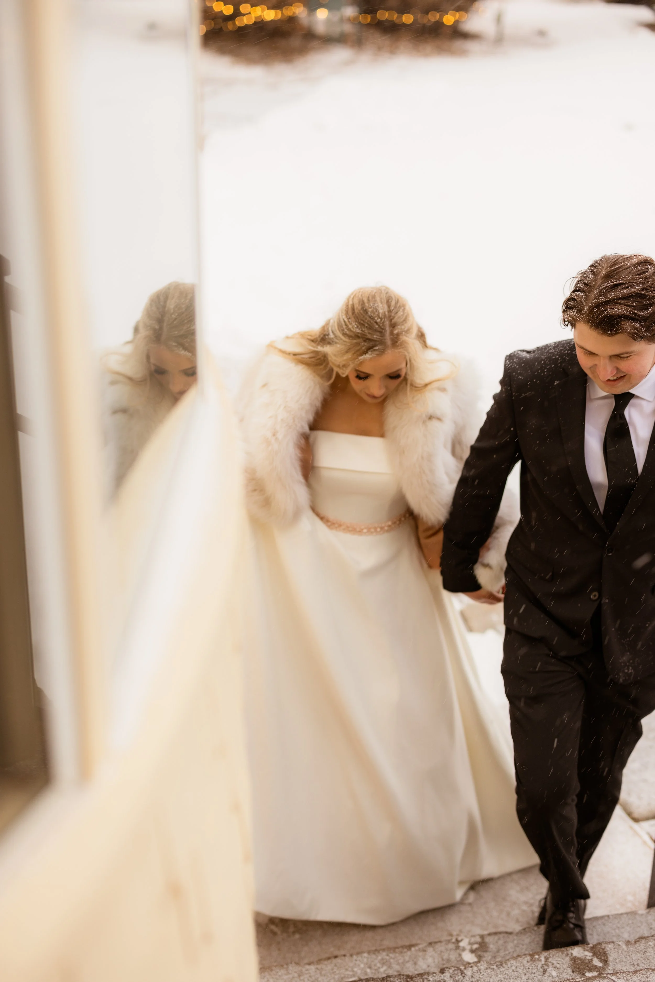 A bride and groom walk hand in hand outside in the snow, the bride wearing a white strapless wedding gown with a fur stole, and the groom in a black tuxedo, smiling.