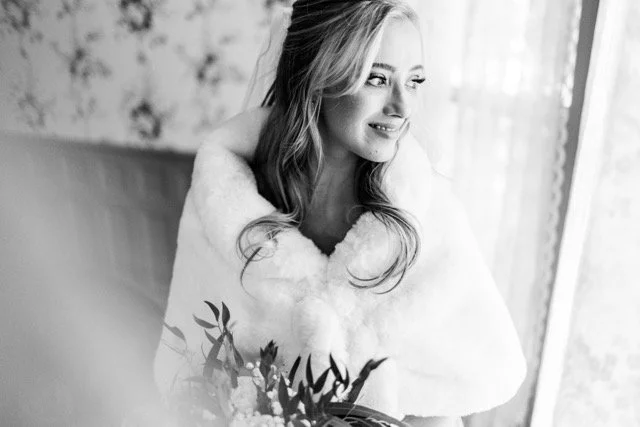 Black and white photo of a woman with long hair, smiling and looking out a window, wearing a fluffy white coat and holding a bouquet of flowers.