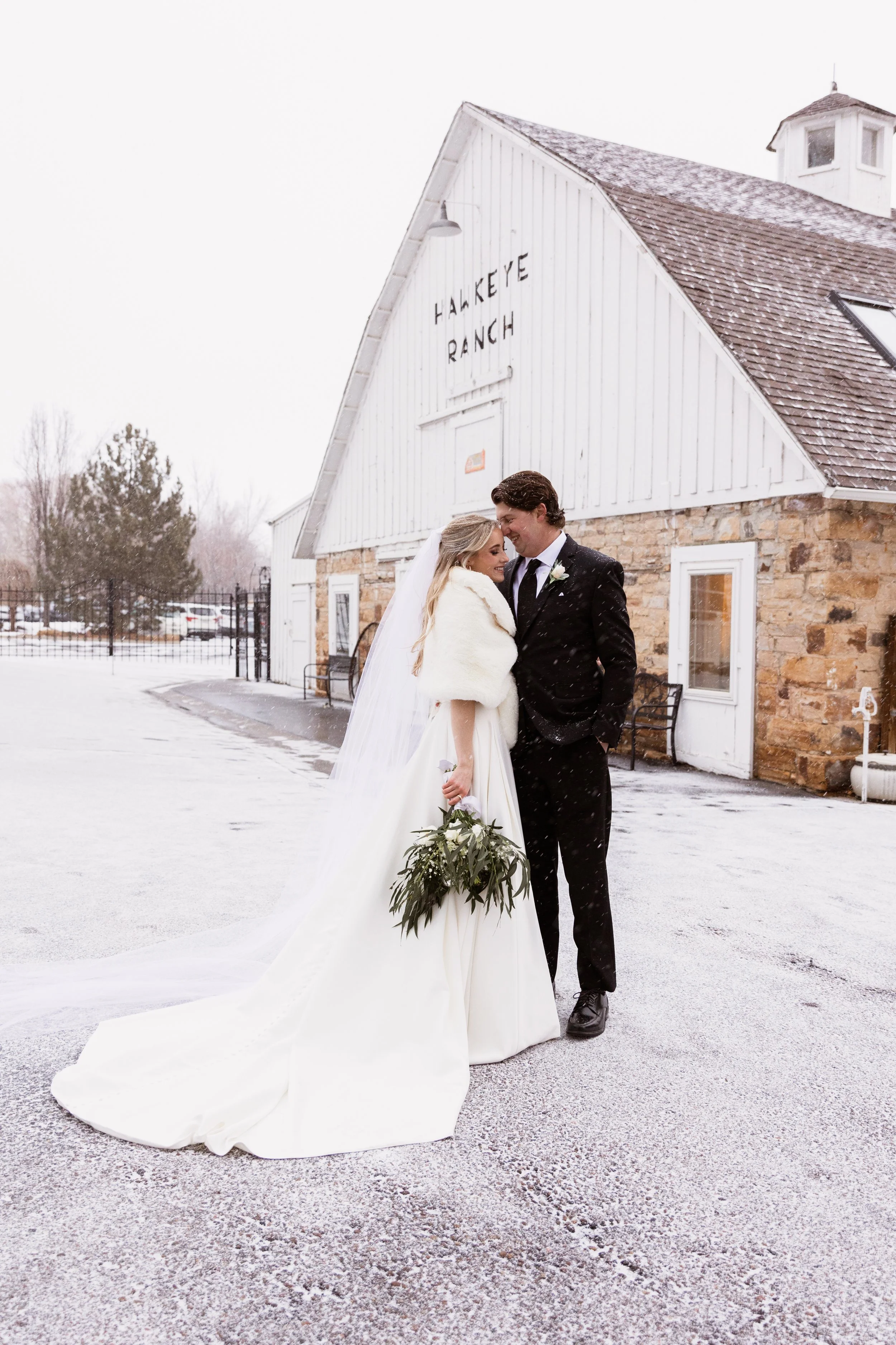 Bride and groom in wedding attire standing close together outdoors in front of Hakkeye Ranch, with snow on the ground and falling snowflakes.