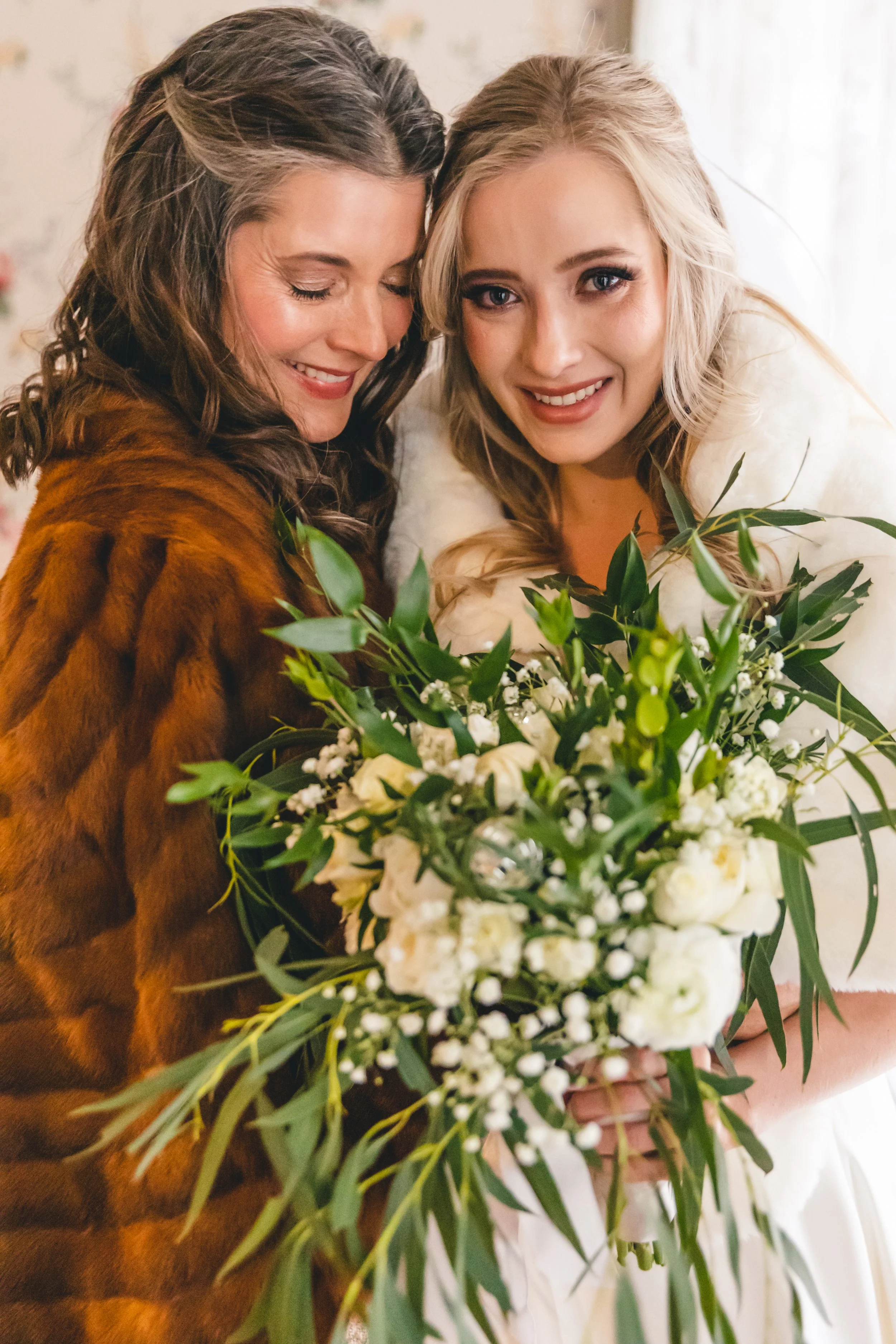Two women, one with dark hair and the other with blonde hair, smiling and holding a bouquet of white flowers, dressed in elegant attire, possibly at a celebration or event.